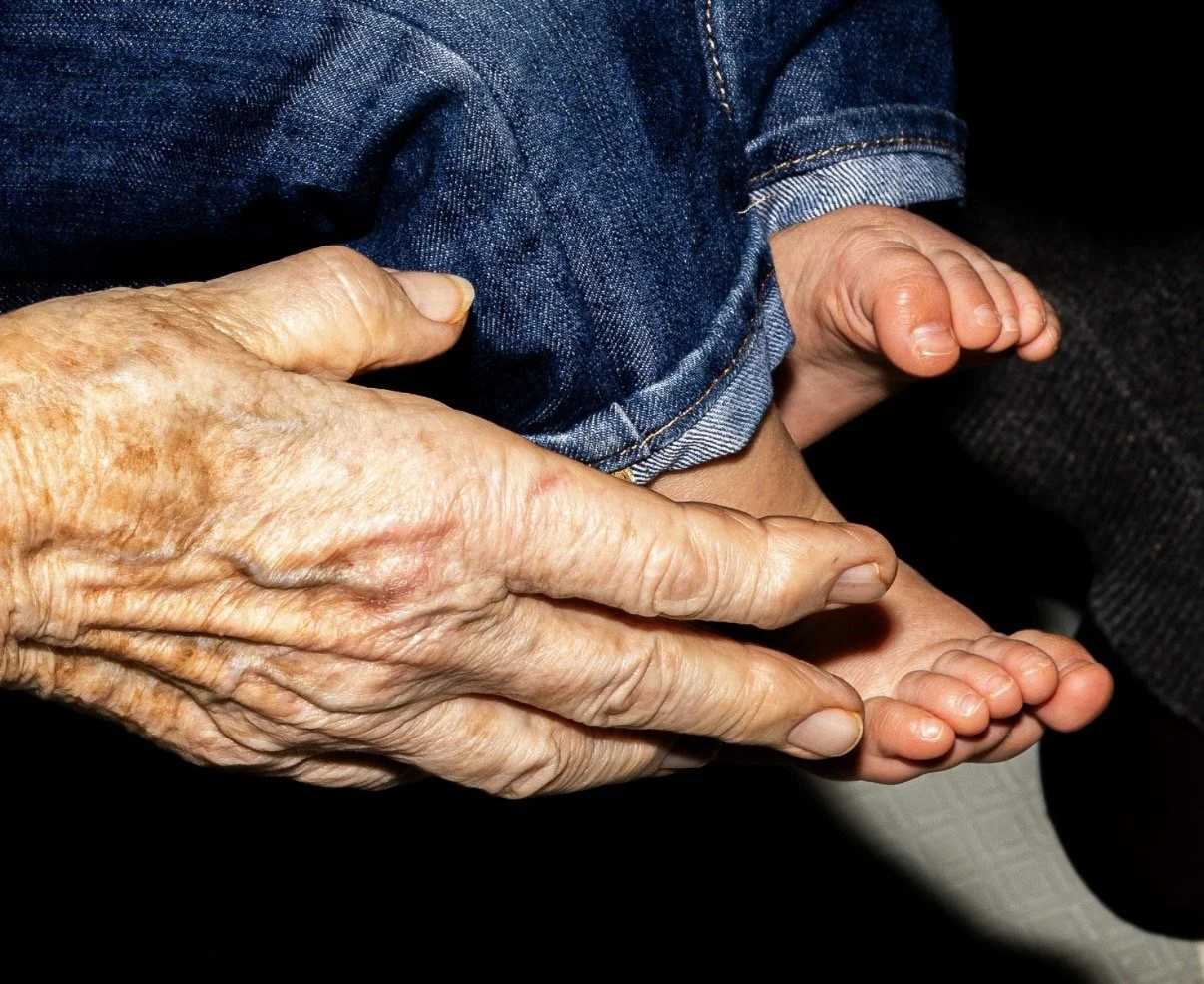 Photographie au flash, d'une main âgée tenant les pieds d'un enfant, toutes deux posées côte à côte.