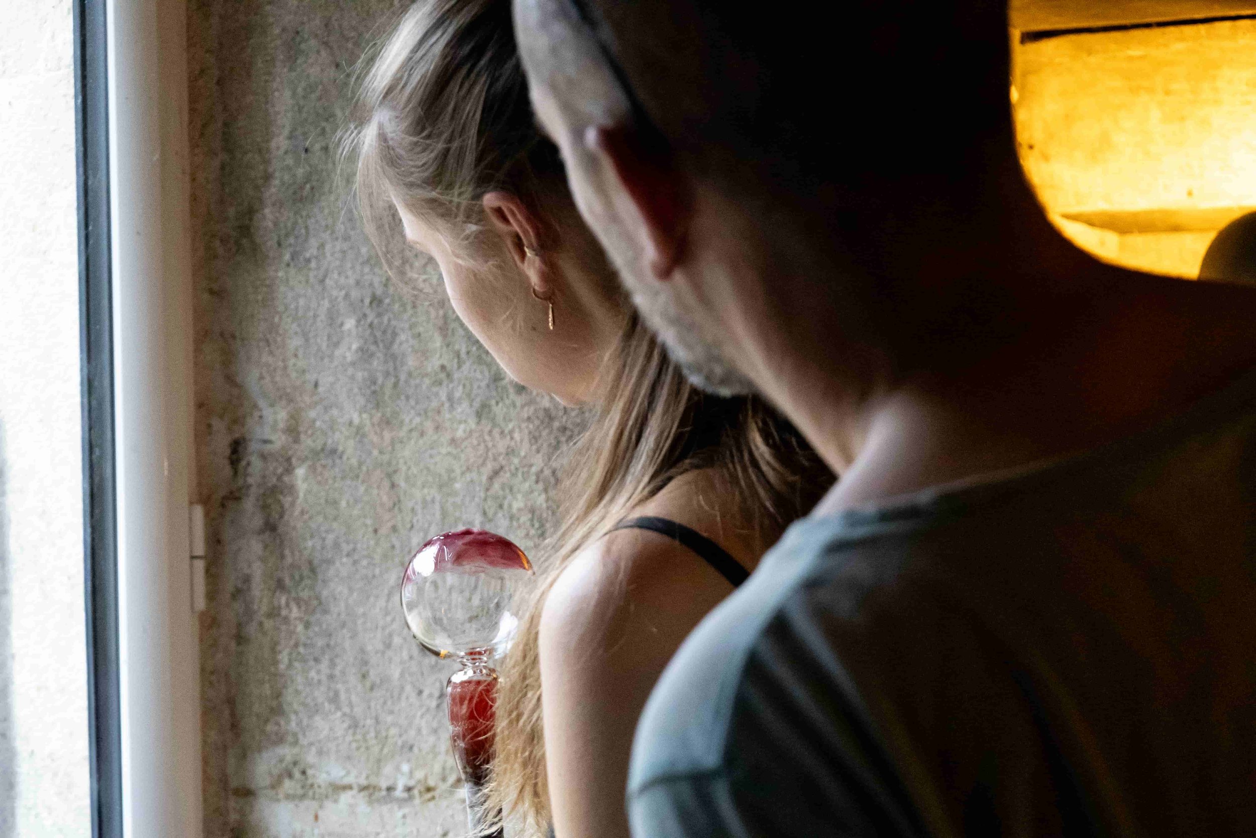 Photographie d'une femme et un homme artistes verriers, se tenant debout à côté d'une fenêtre, regardant un pièce en verre finale, dans un intérieur avec un mur en béton brut et une lampe à lumière chaude.