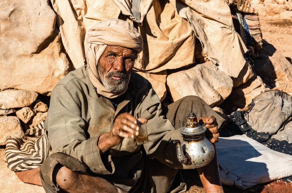 Photographie d'un homme âgé berbère avec une barbe grise, portant un turban beige, assis devant des rochers, tenant une théière en métal et une tasse en verre contenant du thé, dans un environnement désertique dans l'Atlas Marocain. 