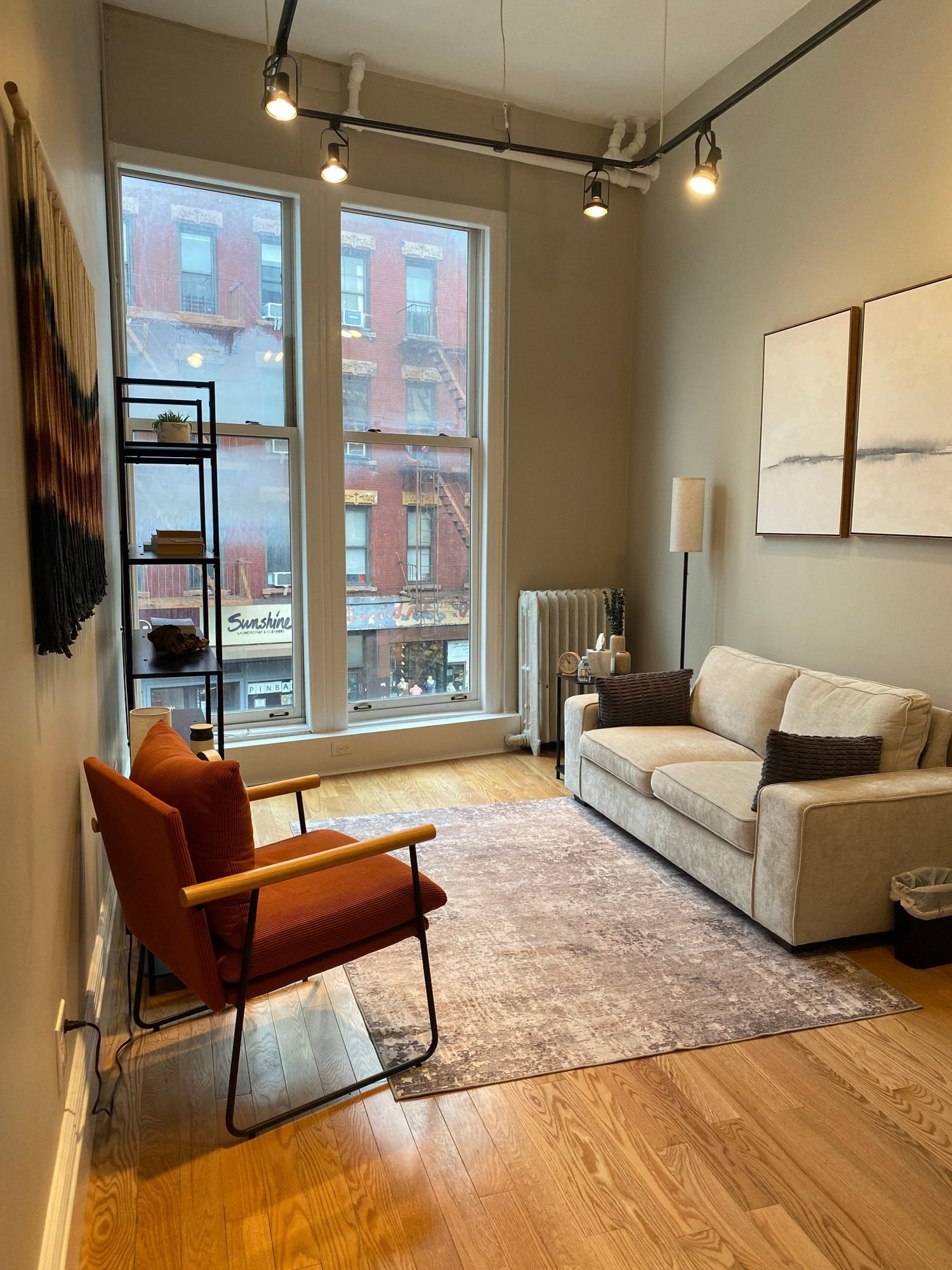 A cozy living room with a beige sofa, orange armchair, a floor lamp, a framed artwork on the wall, large windows, and wooden flooring.