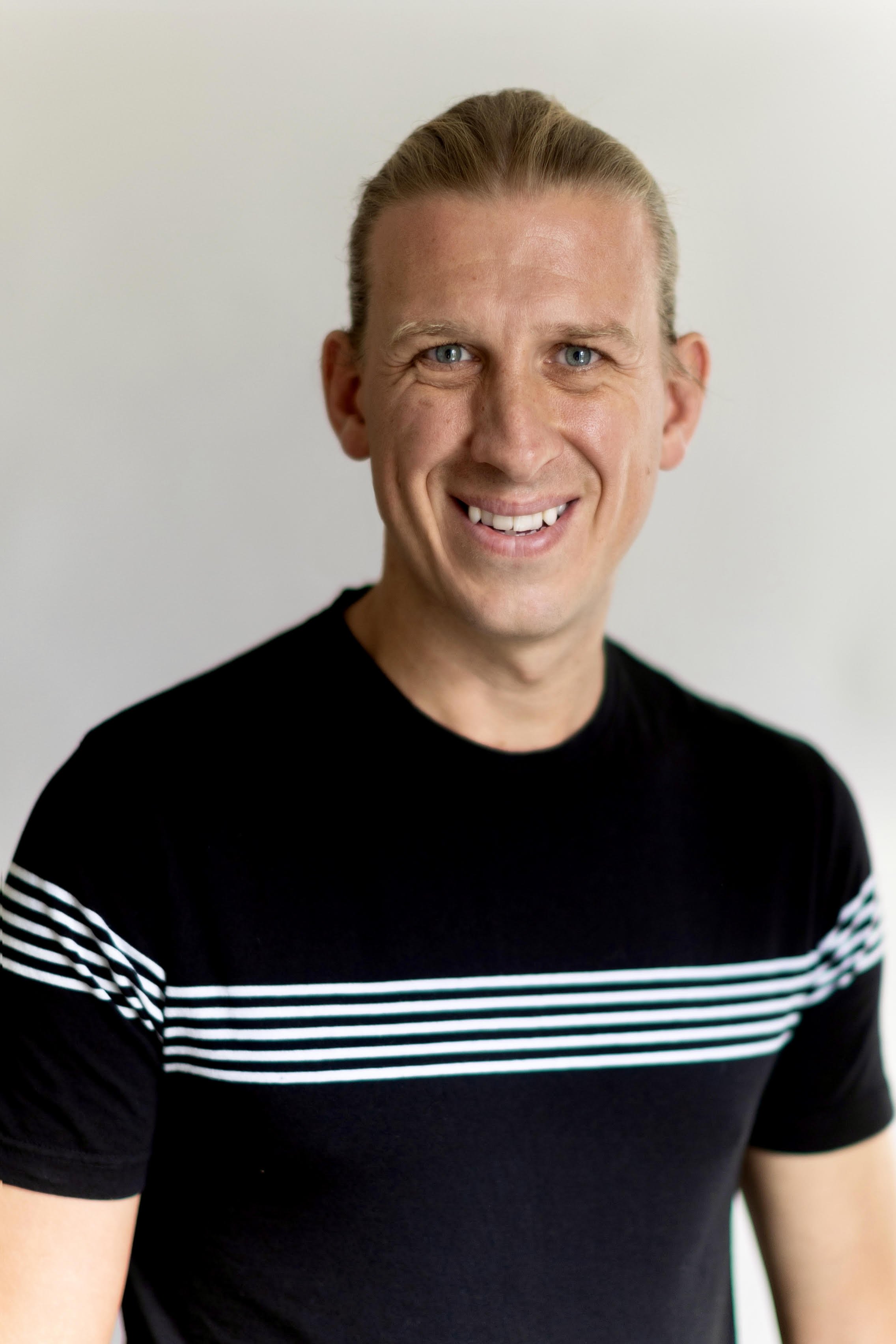 A smiling man with blond hair, blue eyes, and fair skin, wearing a black shirt with white horizontal stripes.