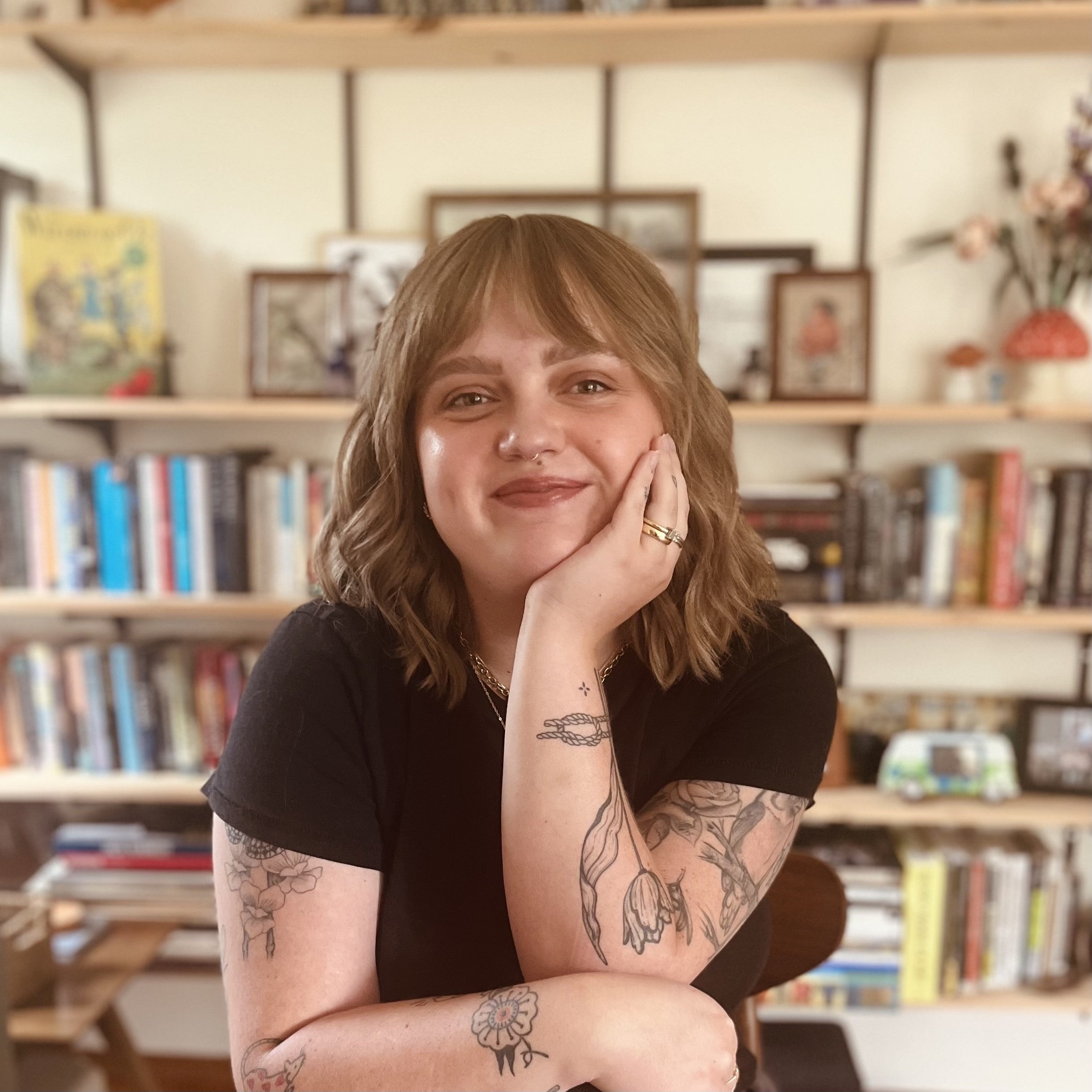 A portrait of Kyla, her head is resting on her hand sitting in front of the bookshelf in her home