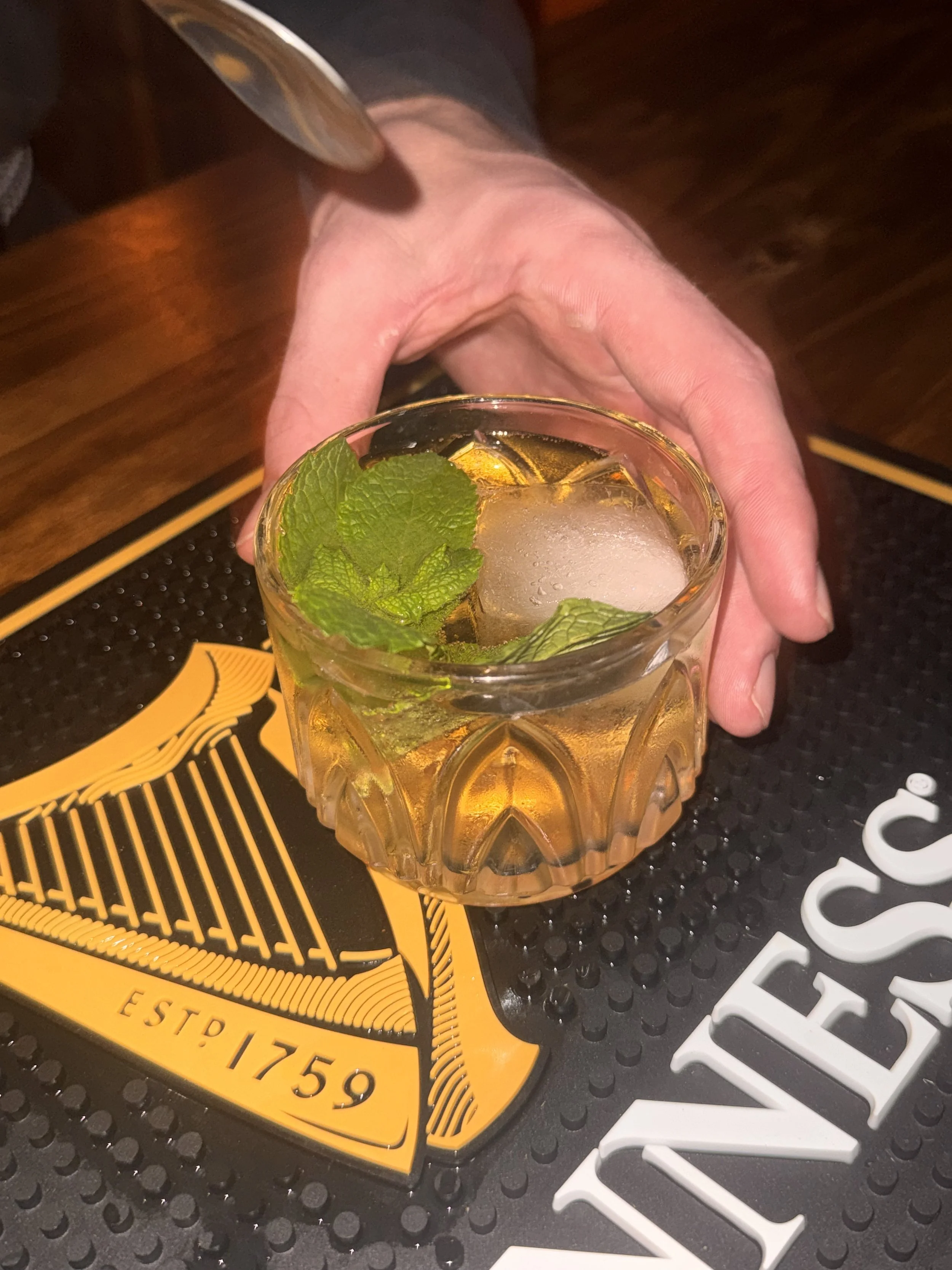 A hand extends over a glass of whiskey with ice and mint leaves, placed on a bar mat with Guinness branding.