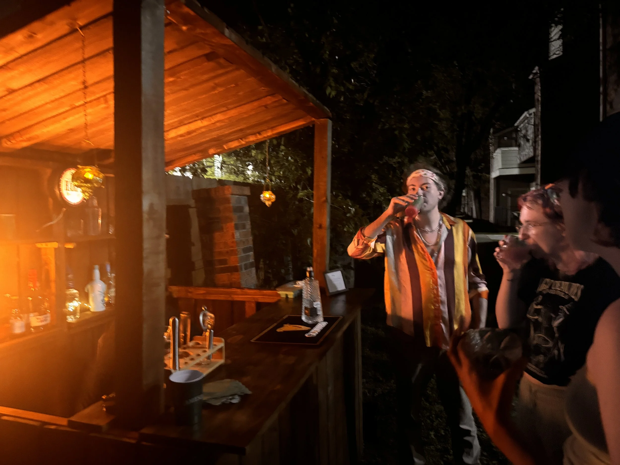Three people at an outdoor bar at night, with one person drinking from a glass and two others with drinks in their hands, a wooden bar structure, bottles on shelf, and warm lighting.