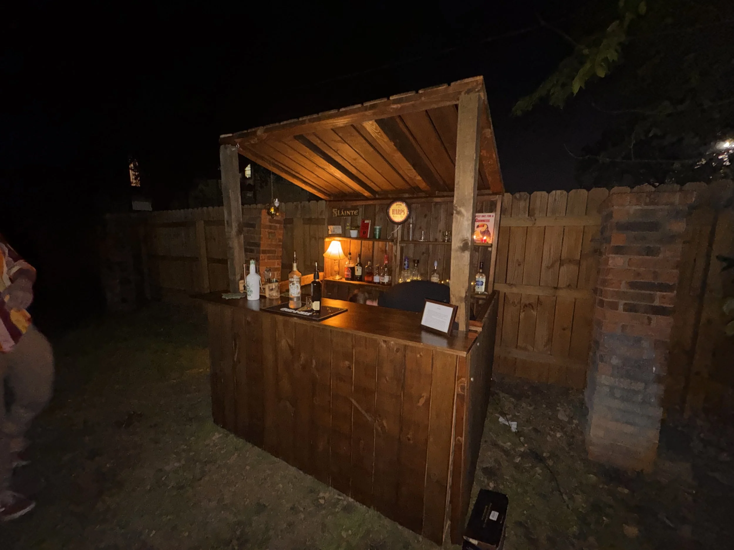 A wooden outdoor bar with bottles and glasses, illuminated by a small lamp, set against a wooden fence at night.