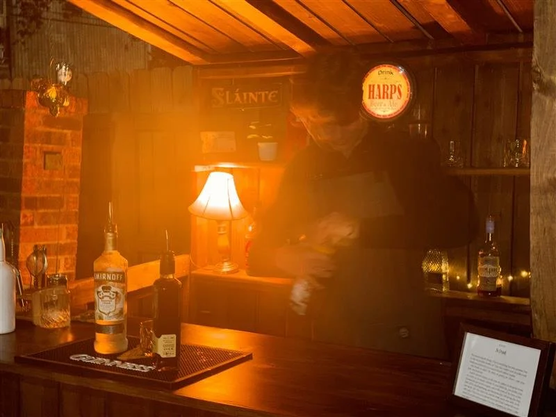 A bar with a wooden counter, bottles of alcohol, a small lamp, and a person holding a drink in a dimly lit, cozy setting.