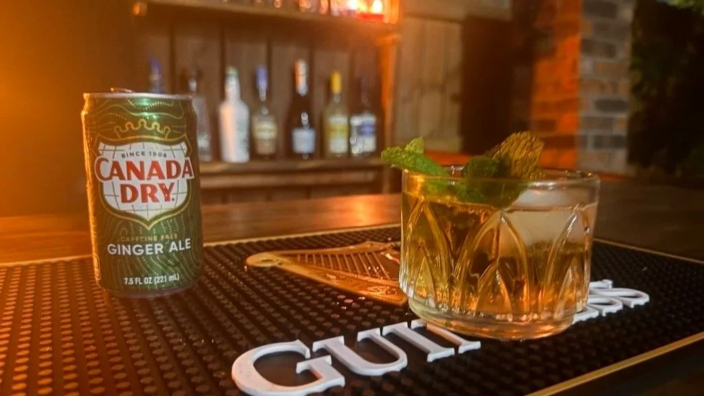 A can of Canada Dry Ginger Ale and a glass of ginger ale with mint leaves on a bar counter.