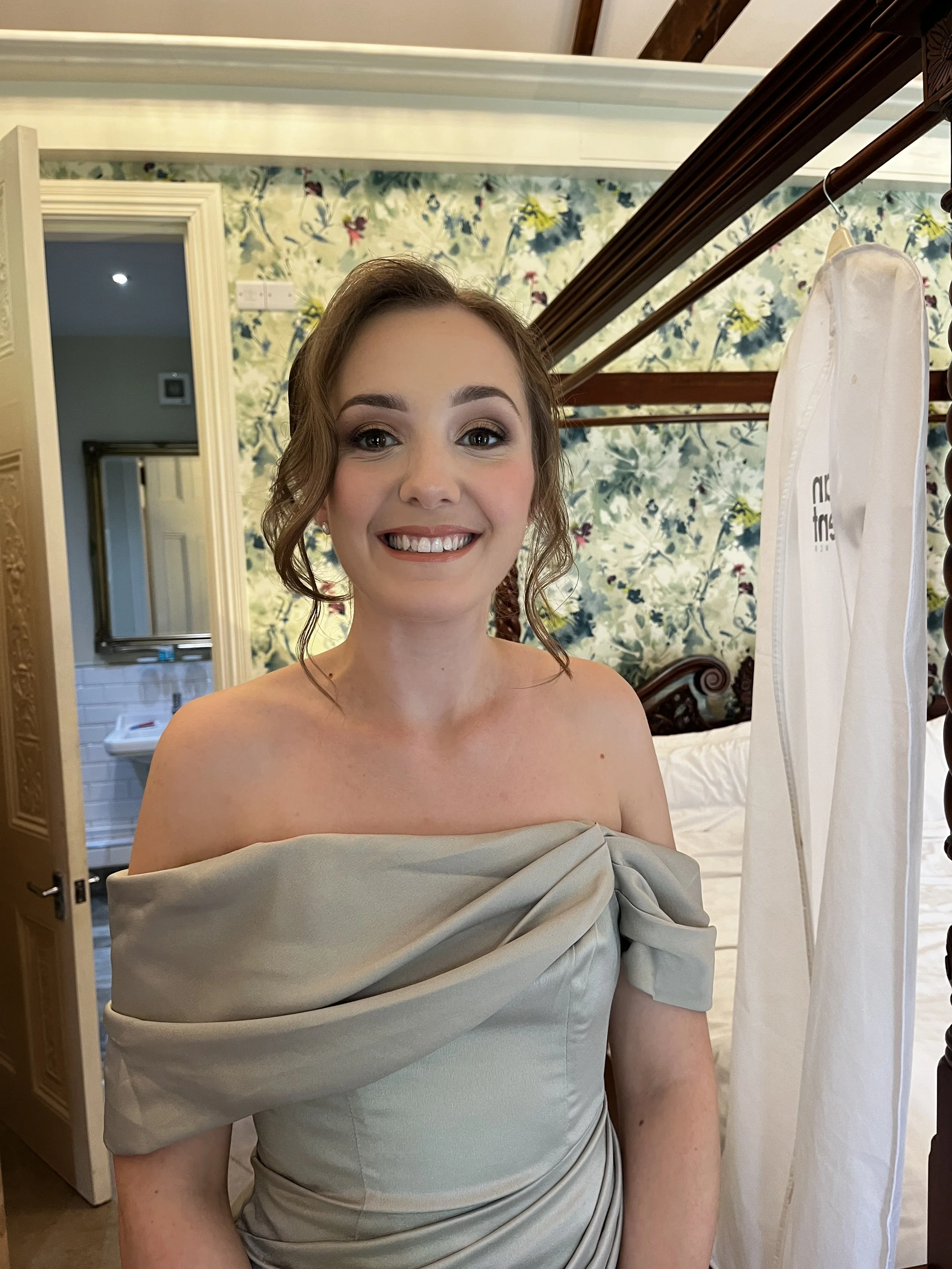 A woman with brown hair styled in loose waves, wearing makeup, smiling, dressed in an off-the-shoulder light beige dress with draped fabric, standing indoors in front of a floral wallpapered wall.
