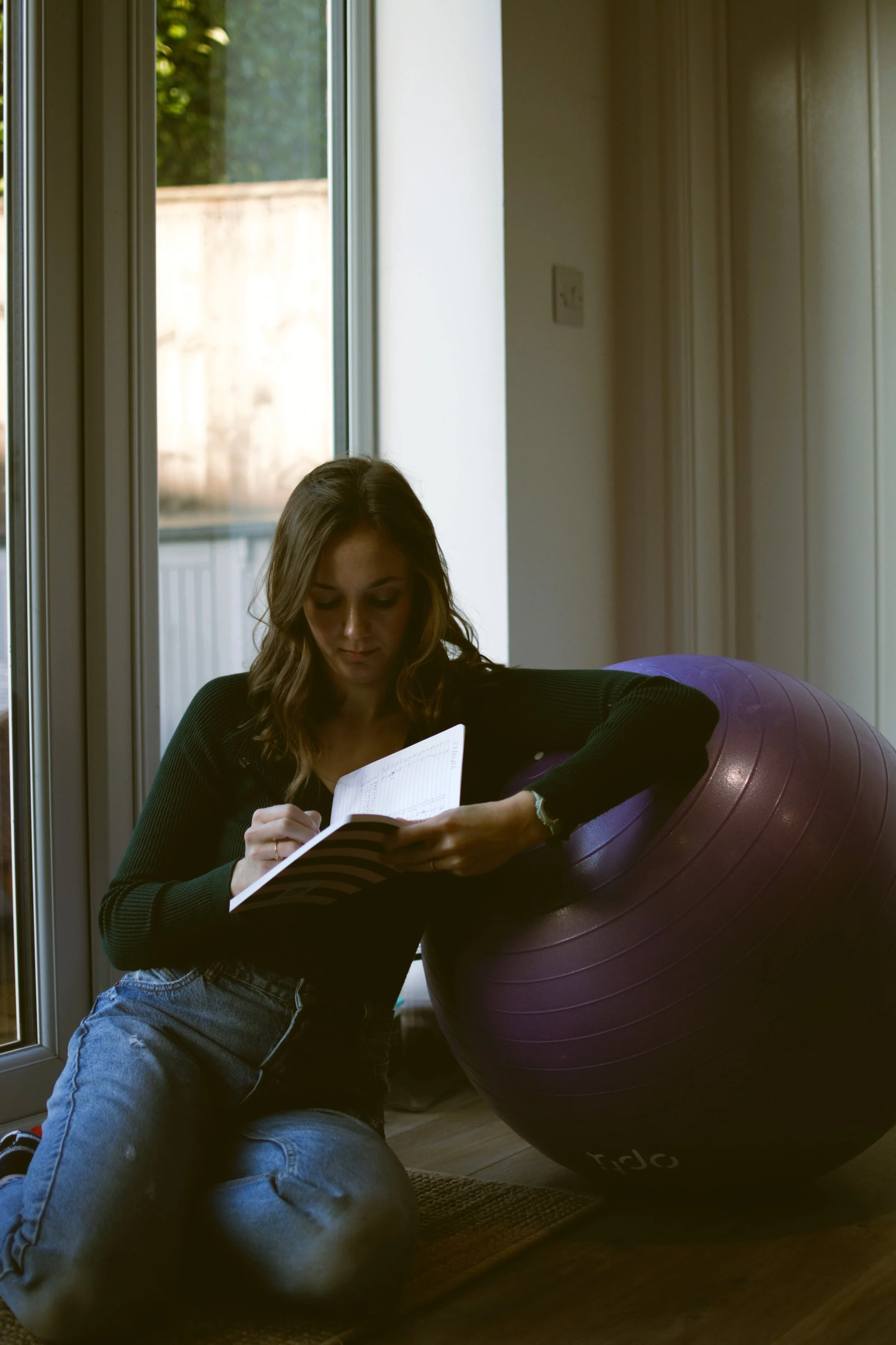 A young woman sitting on the floor next to a large purple exercise ball, writing in a notebook, near a window letting in natural light.
