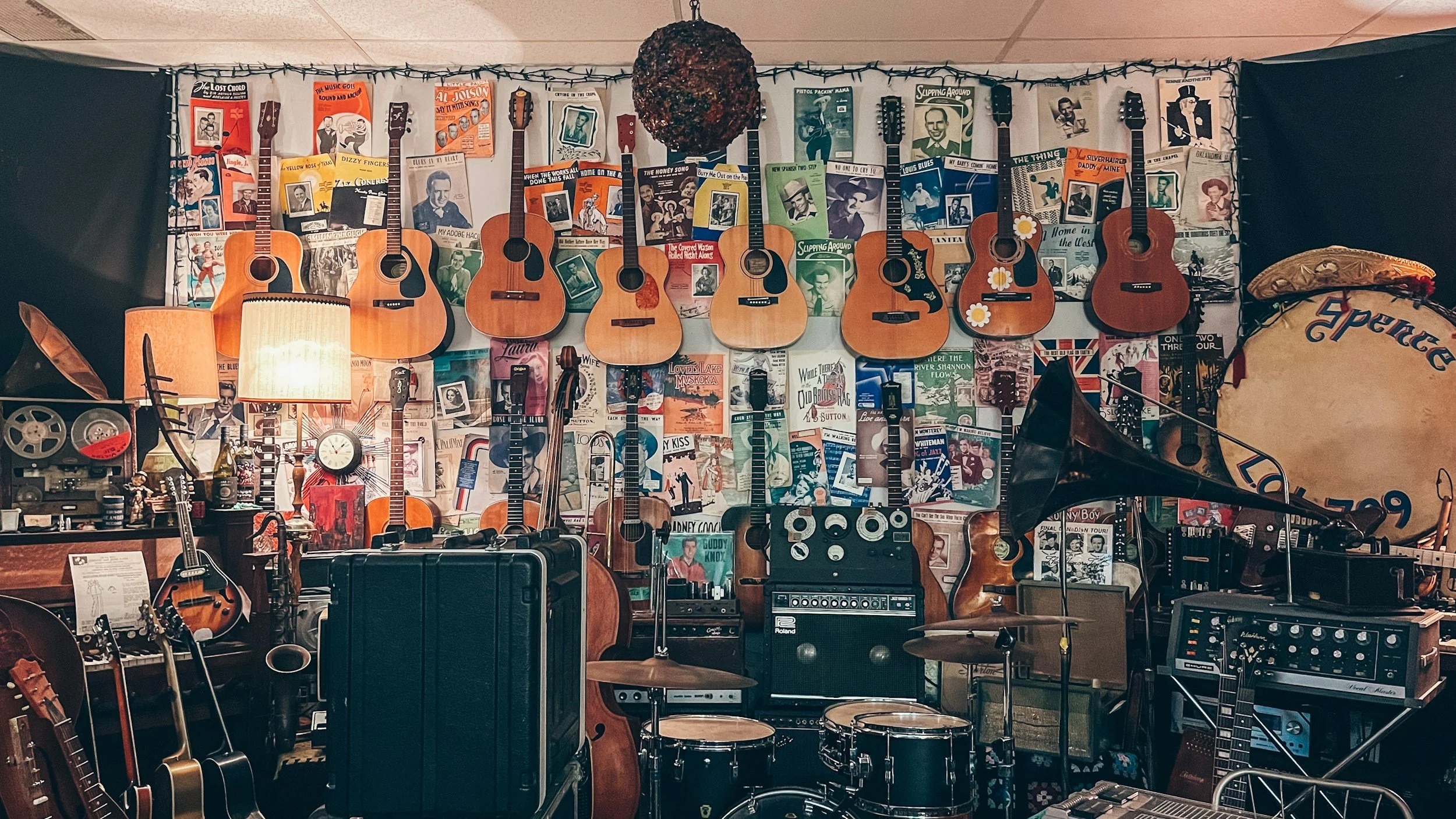 A music room with guitars hanging on the wall, a large drum, and various musical equipment including amplifiers, microphones, and a record player, decorated with posters and string lights.