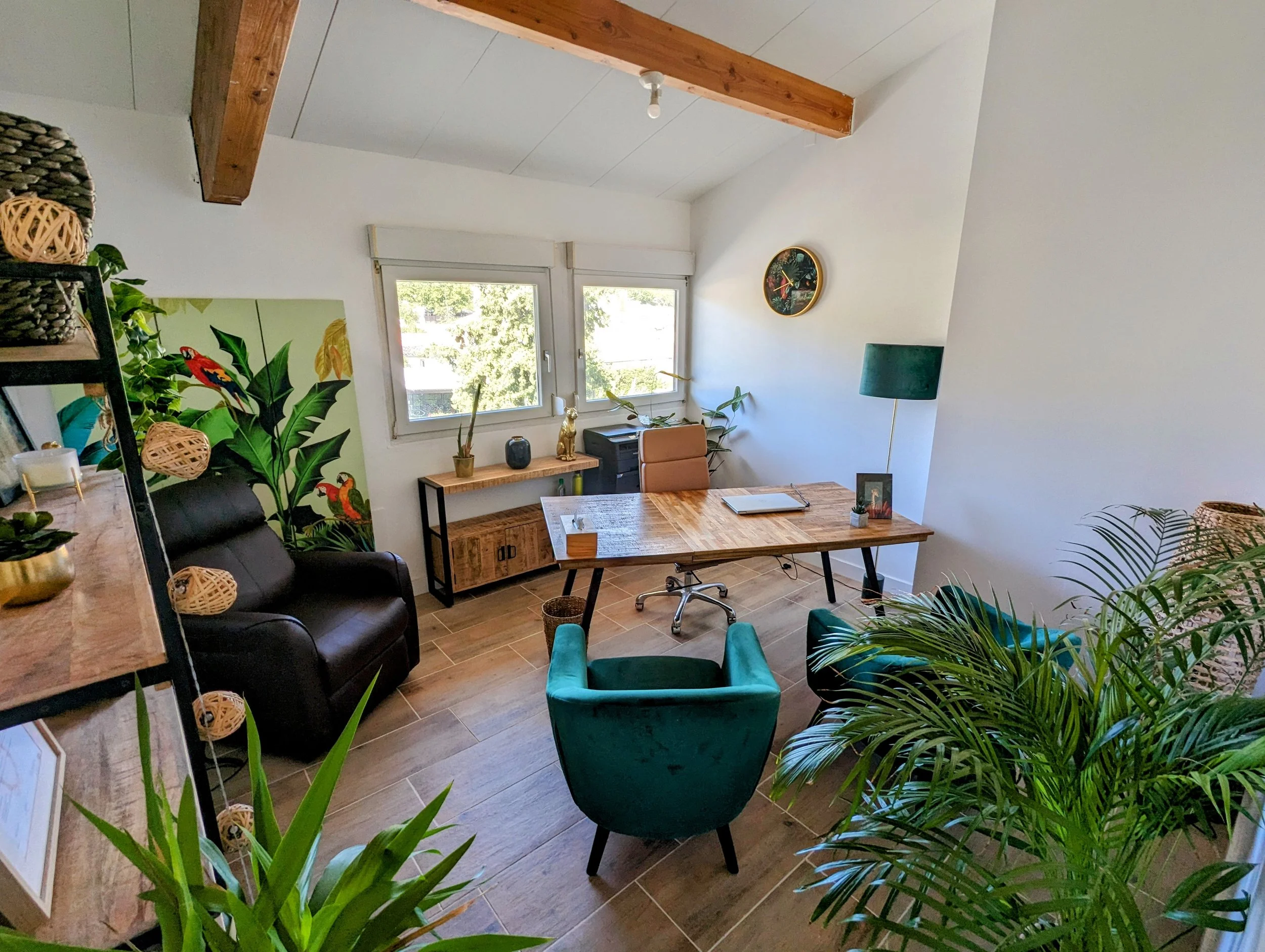 Bureau avec un bureau en bois, un fauteuil vert, un fauteuil noir, une lampe à pied, des plantes vertes, un mural végétal avec des perroquets, et une étagère avec décorations.