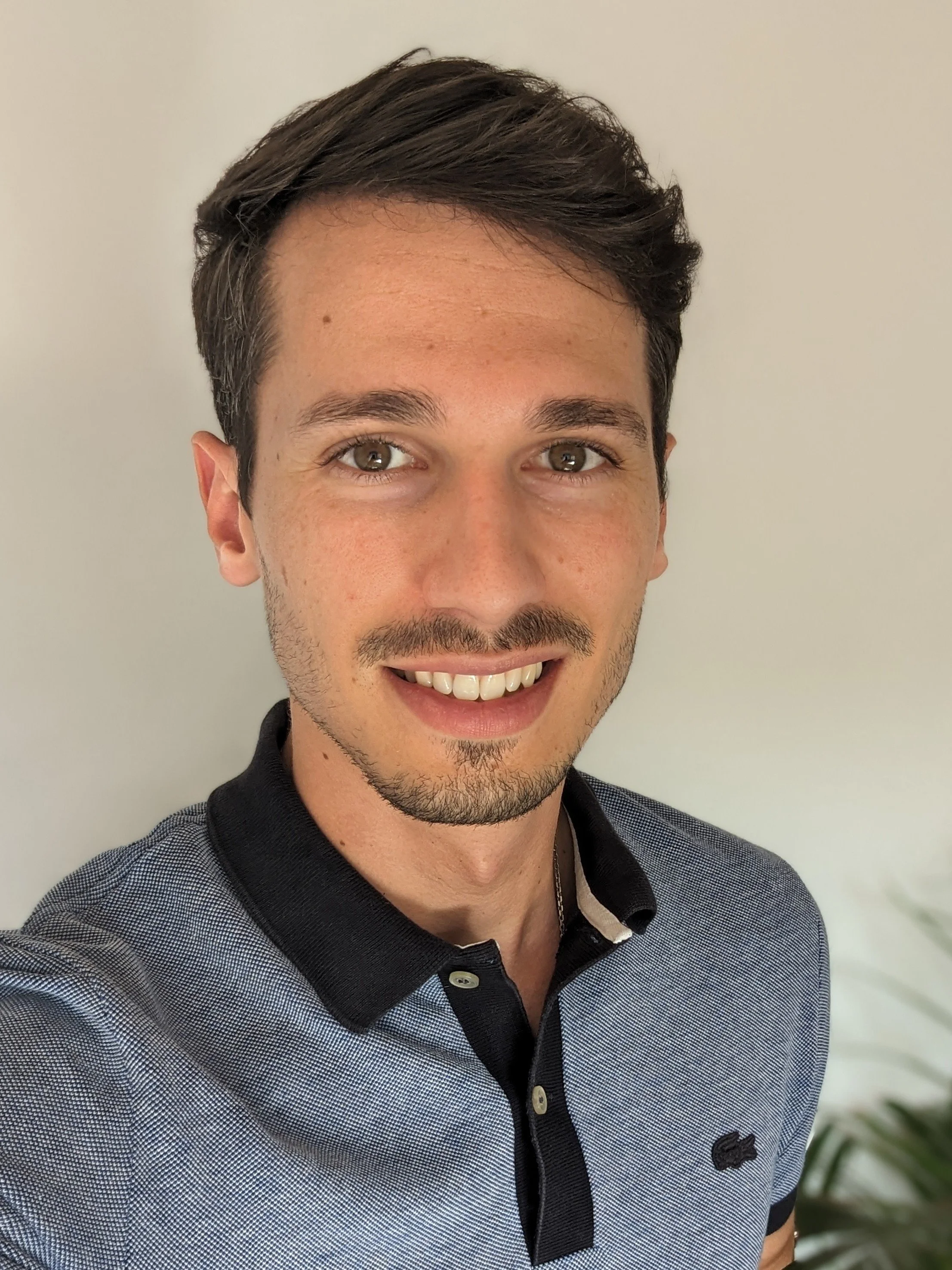 Un jeune homme souriant avec des cheveux bruns, portant une chemise à col noir avec un motif en petits points bleus, se tenant devant un mur blanc.
