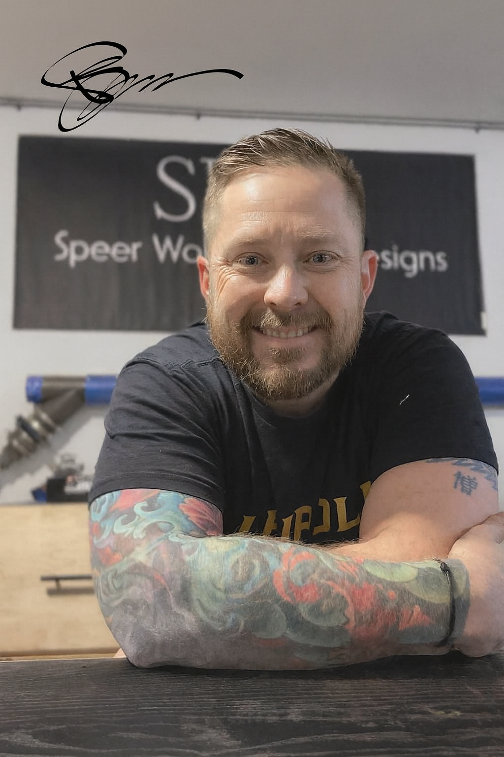 Man with tattooed arms smiling, leaning on a black table in a gym, with a sign in the background that reads 'Speer Web Designs'.