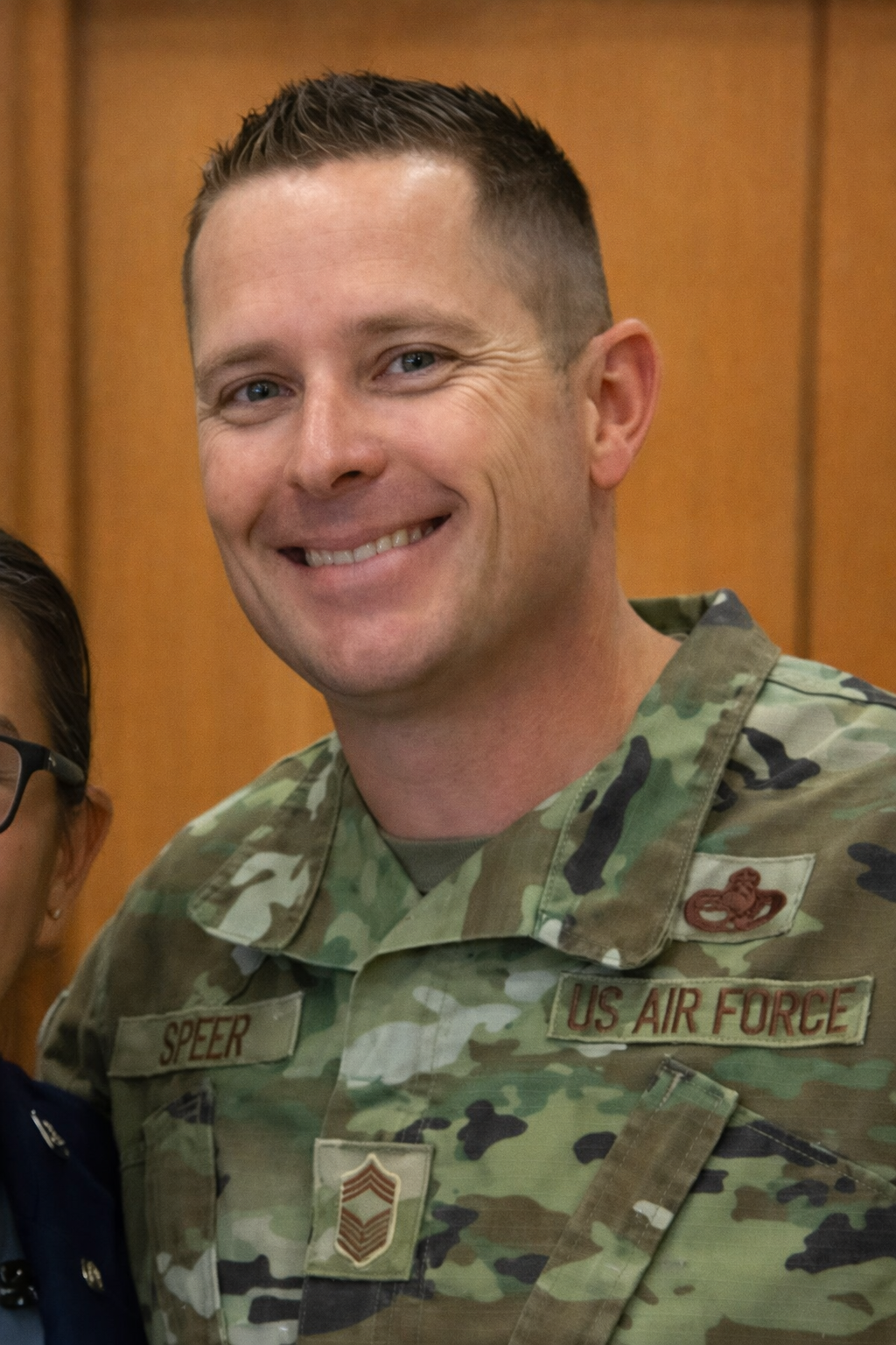 Male soldier in camouflage uniform smiling, with a U.S. Air Force patch and rank insignia, standing in a wood-paneled room.