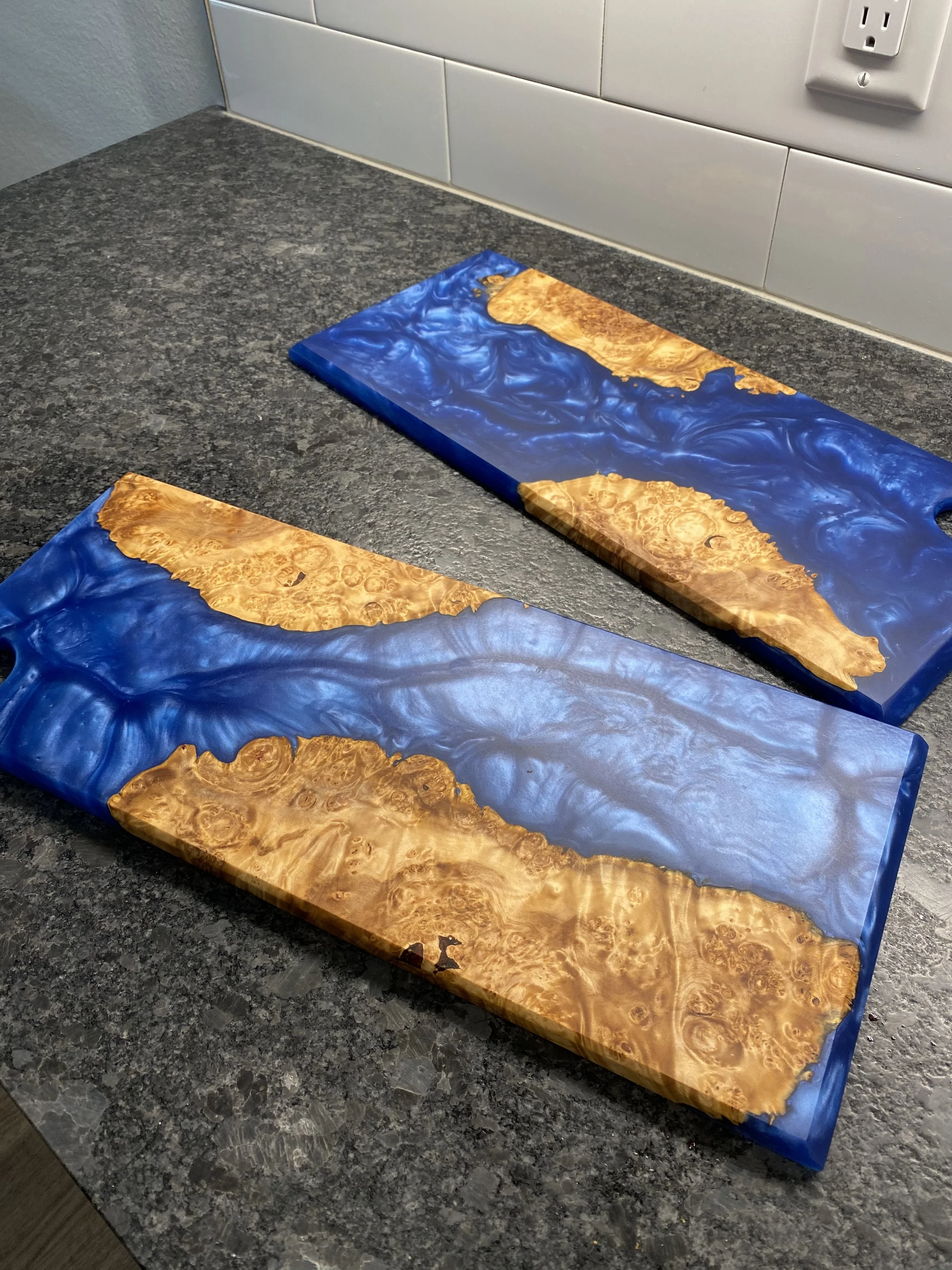 Two rectangular wooden cutting boards with blue resin inlays, resting on a graystone countertop.