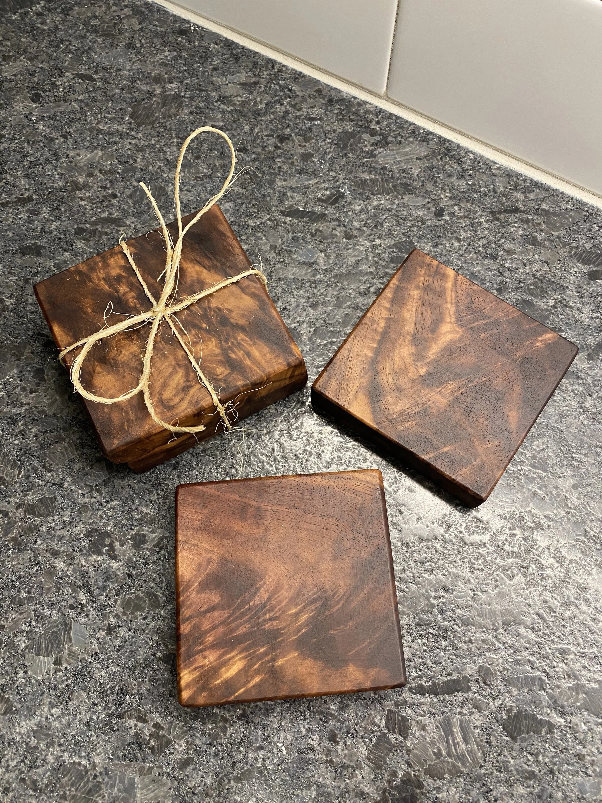 Three wooden coaster or small tray items with a dark, polished finish on a speckled gray countertop. One of the items is wrapped with twine forming a bow.