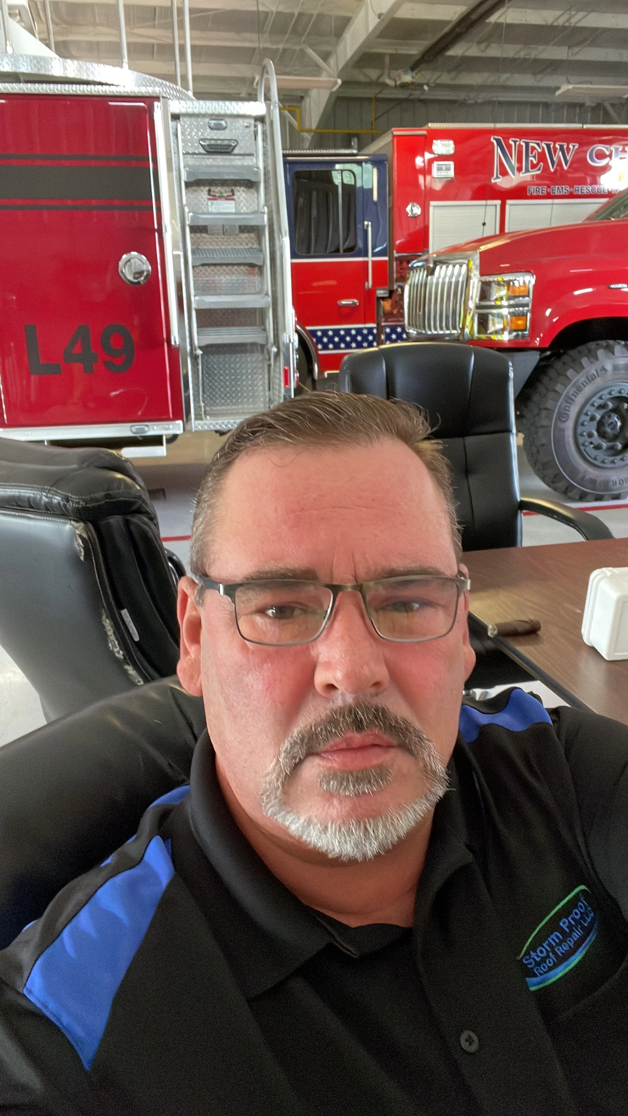 A man with glasses and a gray beard taking a selfie in front of fire trucks inside a fire station.