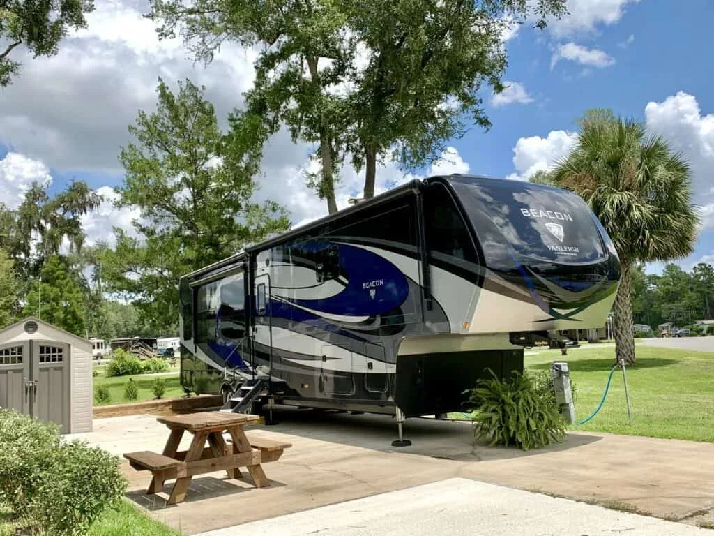 A large black and blue RV trailer parked on a concrete pad in a grassy area with trees and a bright sky with clouds.