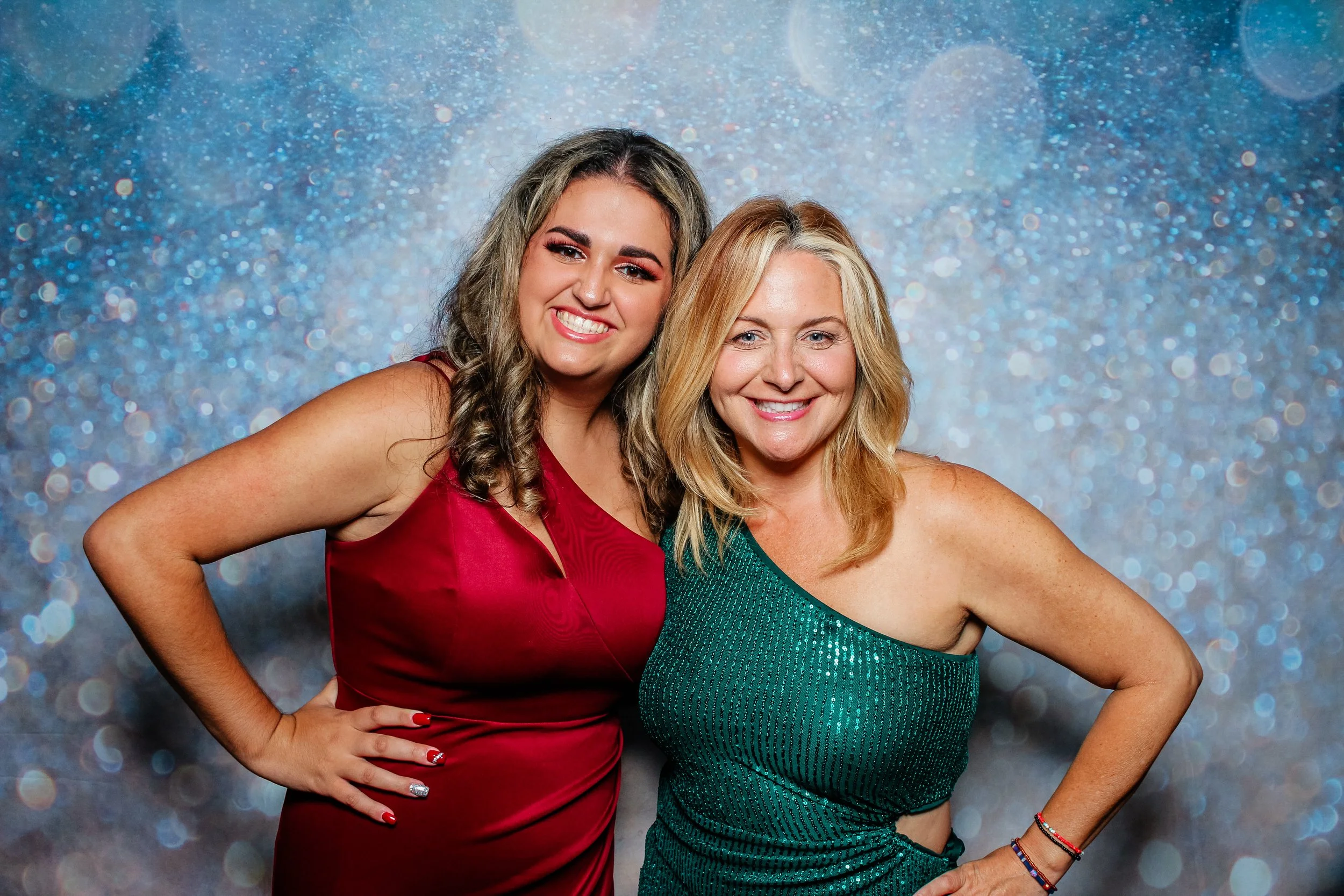 Two women in dresses posing and smiling, standing close together against a glittery blue background.