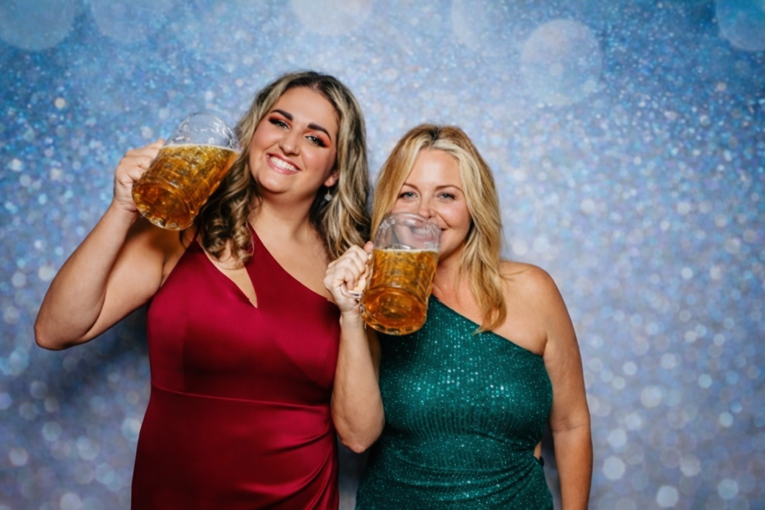 Two women smiling and holding large mugs of beer at a festive event, with a sparkly blue background.