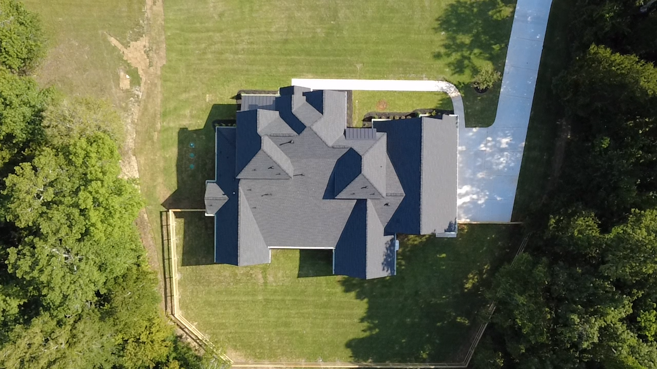 Aerial view of a house with a dark gray roof, surrounded by a green yard with trees and a paved driveway.