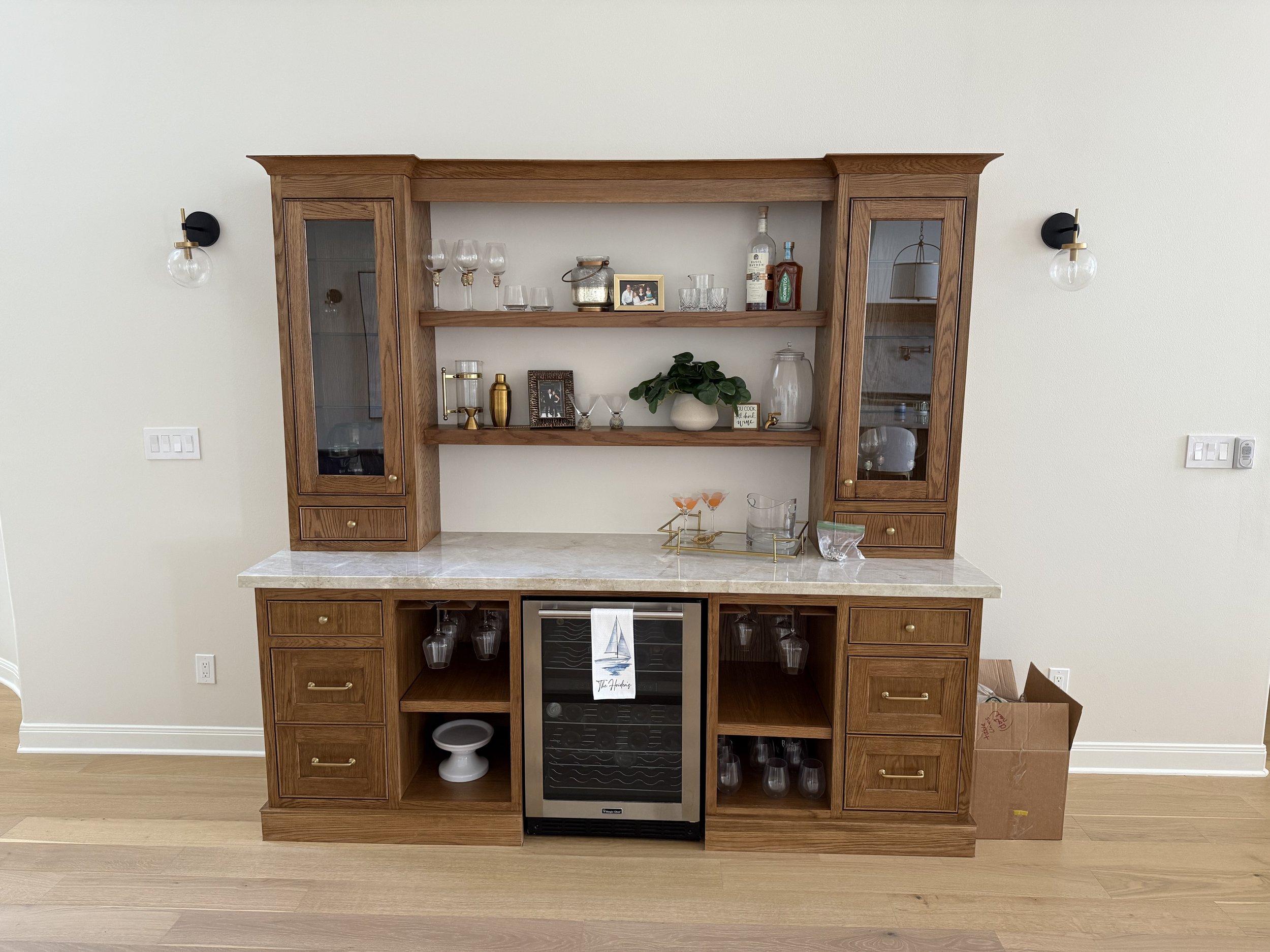 A wooden bar cabinet with glass doors, shelves with glassware, alcohol bottles, decorative items, and a wine cooler, against a white wall with wall-mounted lights.