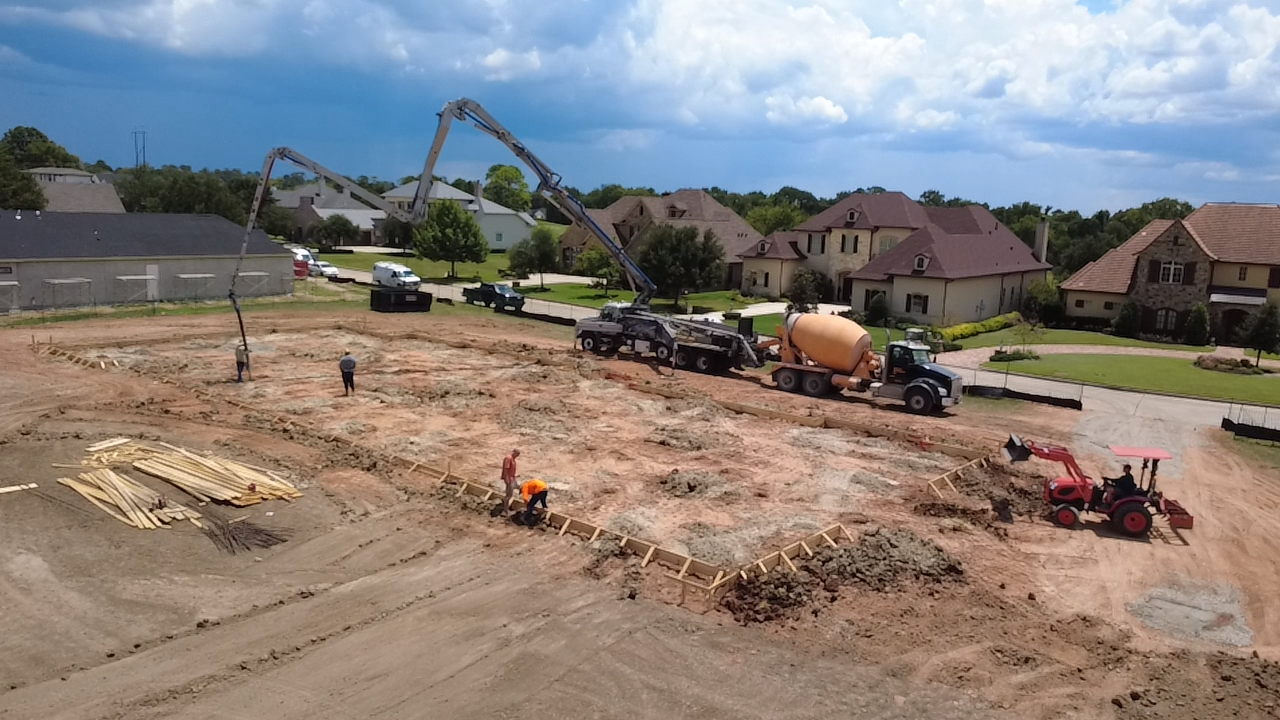 Construction site where workers are preparing the foundation, with a cement mixer truck, concrete pump, and various construction materials and equipment seen.