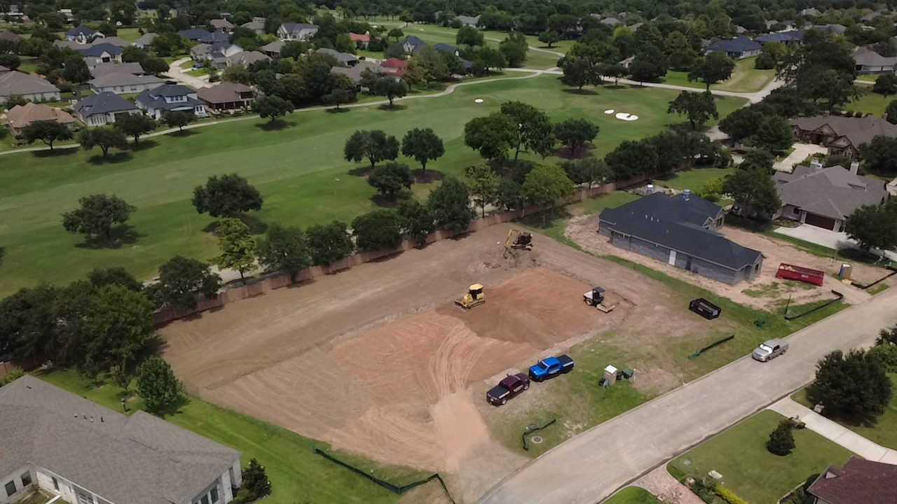 A residential neighborhood with a large green golf course and houses surrounding it. In the foreground, there is a construction site with dirt, construction vehicles, and parked cars.