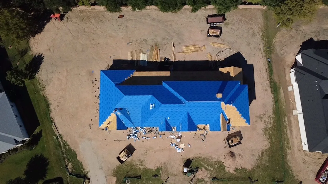 Aerial view of a house under construction with a blue roof, surrounded by a sandy yard, construction materials, and tools.