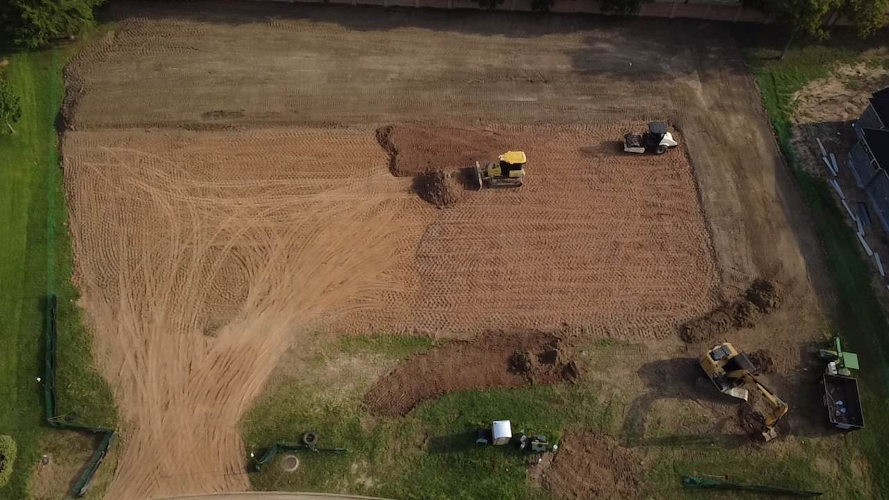 An aerial view of a construction site where land is being excavated and leveled, with heavy machinery such as compactors and excavators working on the area.
