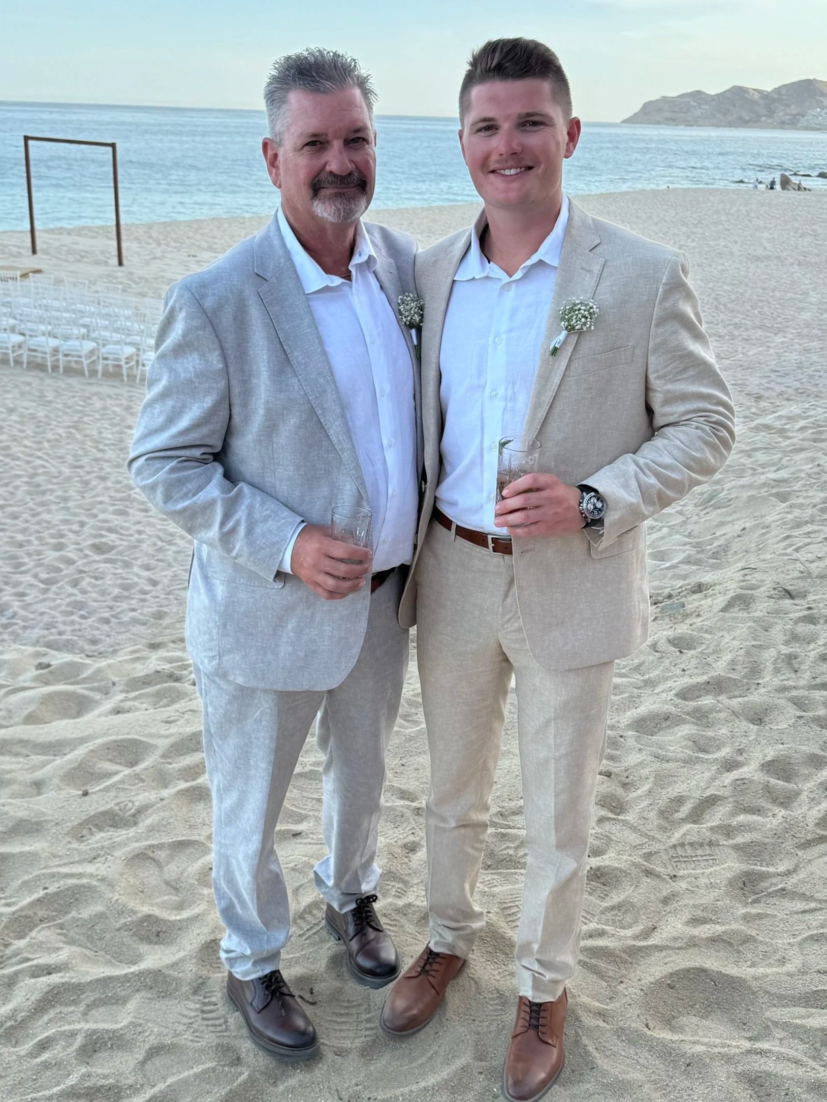 Two men in beige suits standing on a sandy beach near the ocean, holding glasses, with a backdrop of beach chairs, the ocean, and distant mountains.