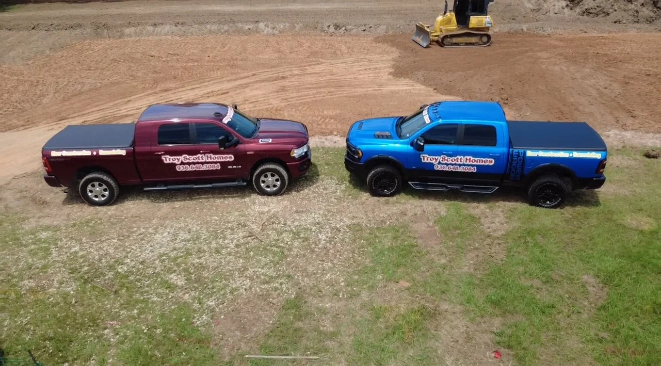 Two pickup trucks, one red and one blue, parked on a grassy area near a construction site; both trucks have advertising decals for Troy Scott Homes. A small yellow bulldozer is working on dirt in the background.