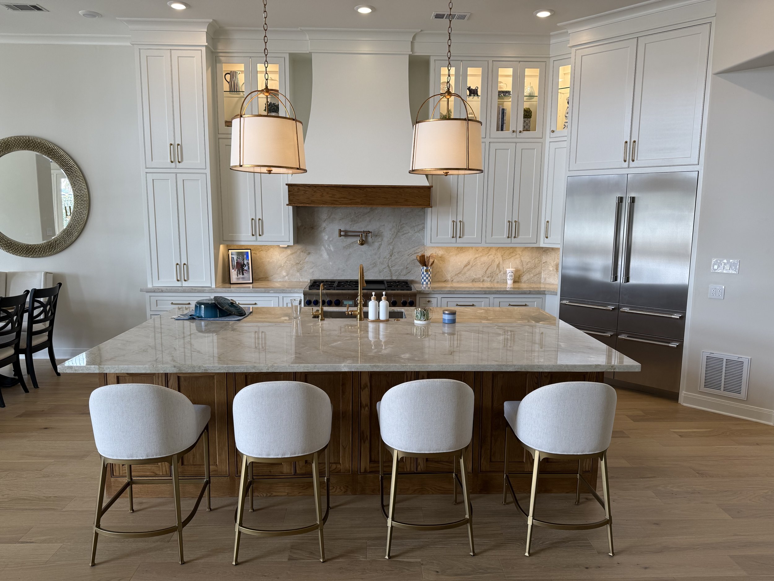 Modern kitchen with cream-colored cabinets, a large marble island, and a stainless steel refrigerator. There are four white chairs at the island, and pendant lights hanging above.