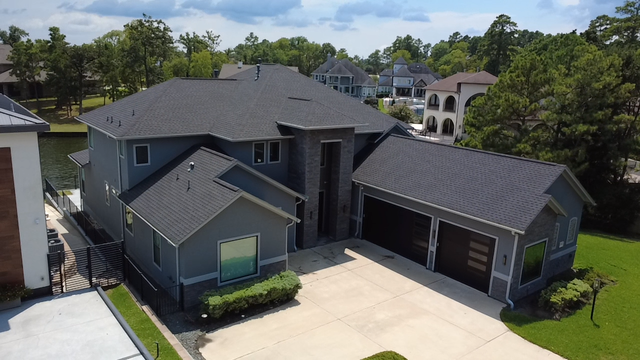 A large modern house with gray siding, multiple roof sections, and two garage doors. Surrounding trees and neighboring houses are visible.