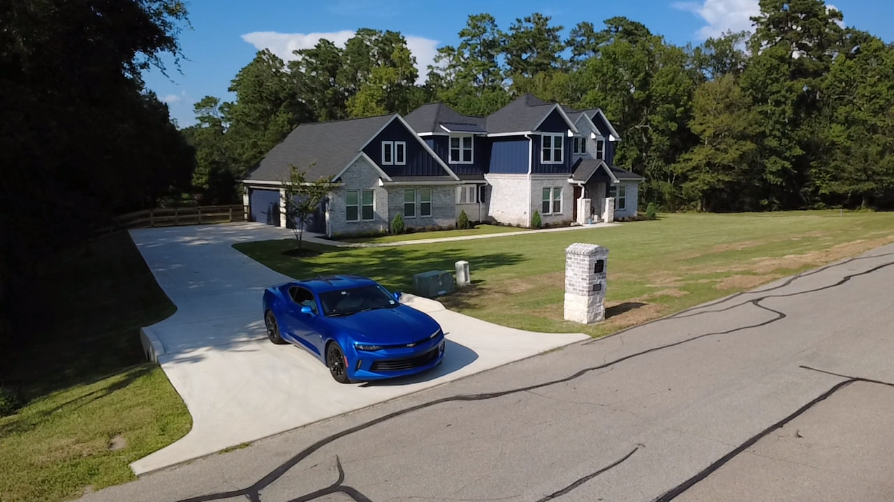 A blue sports car parked in the driveway of a large house with a gray roof and a brick exterior, surrounded by greenery and trees.