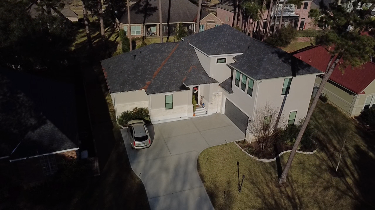 Aerial view of a modern two-story house with a black roof, beige exterior, and attached garage, surrounded by trees and adjacent houses.