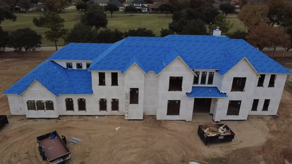 A large house under construction with a blue roof and white exterior walls, surrounded by dirt and construction equipment, with trees and houses in the background.