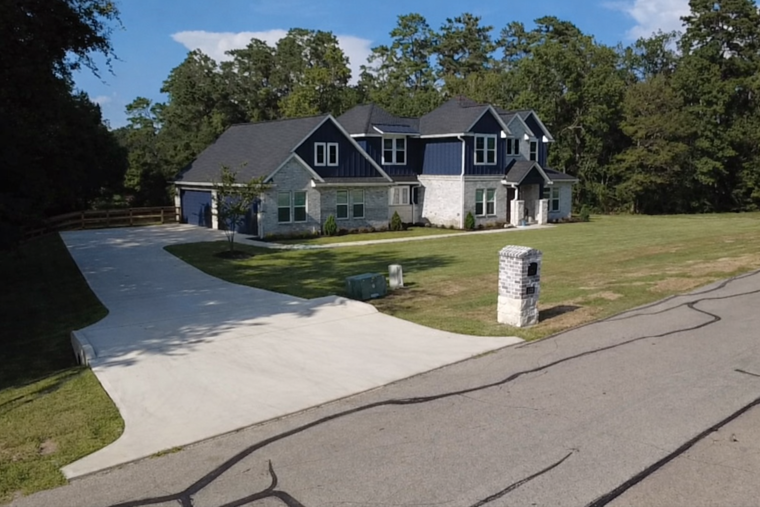 Large two-story house with a stone and dark siding exterior, multiple gables, a driveway, a mailbox, a mailbox, and a well-maintained lawn, surrounded by trees under a blue sky.