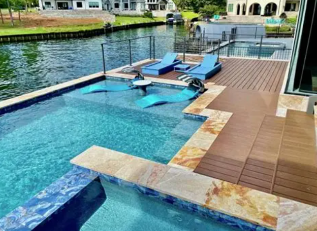 View of a backyard with a swimming pool, water jets, poolside blue lounge chairs, and a wooden deck overlooking a lake.