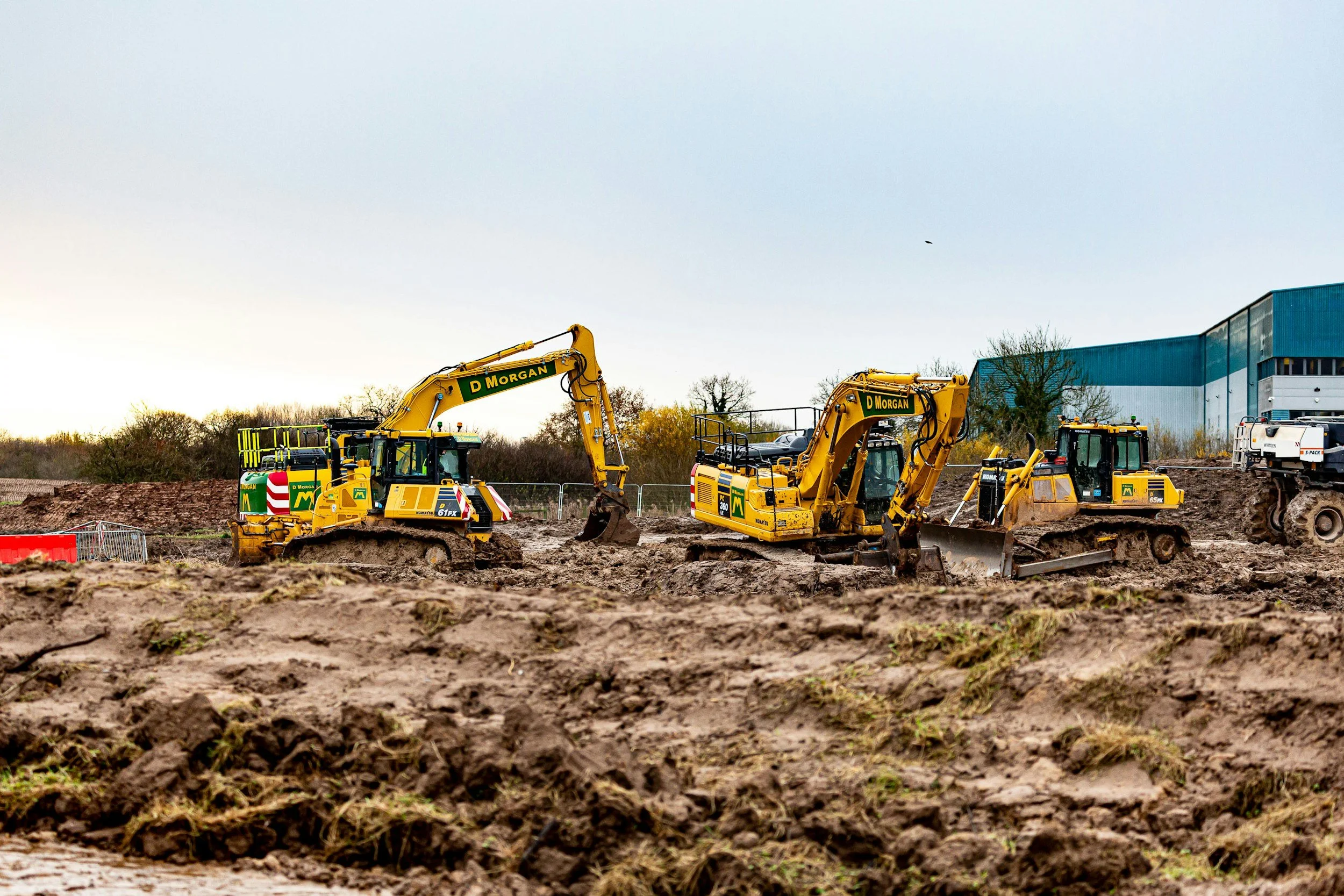 Construction site with three yellow excavators working on dirt, with a large blue building in the background.
