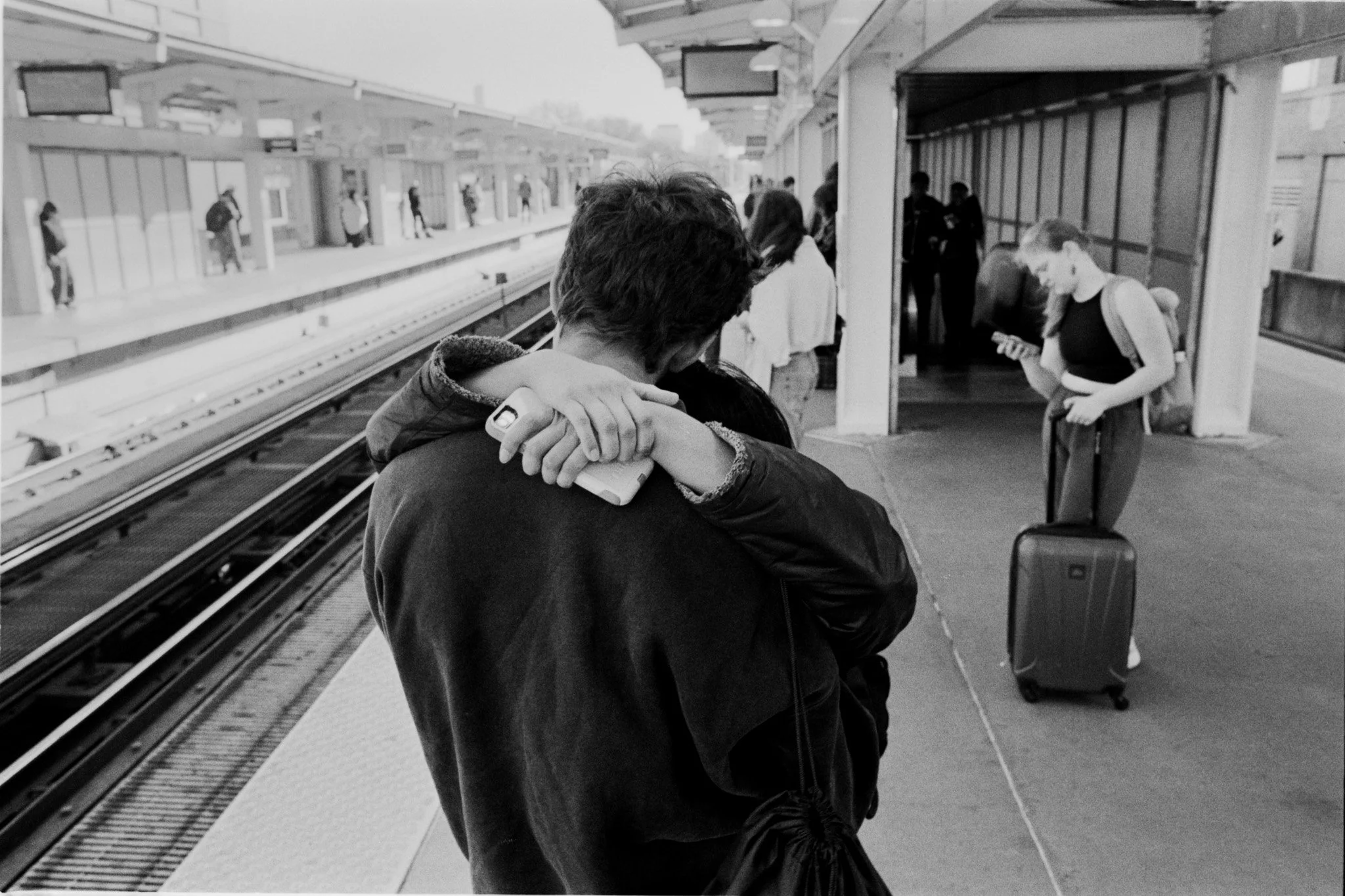 A couple hugging on a train station platform, with people waiting and a woman looking at her phone with a rolling suitcase nearby.