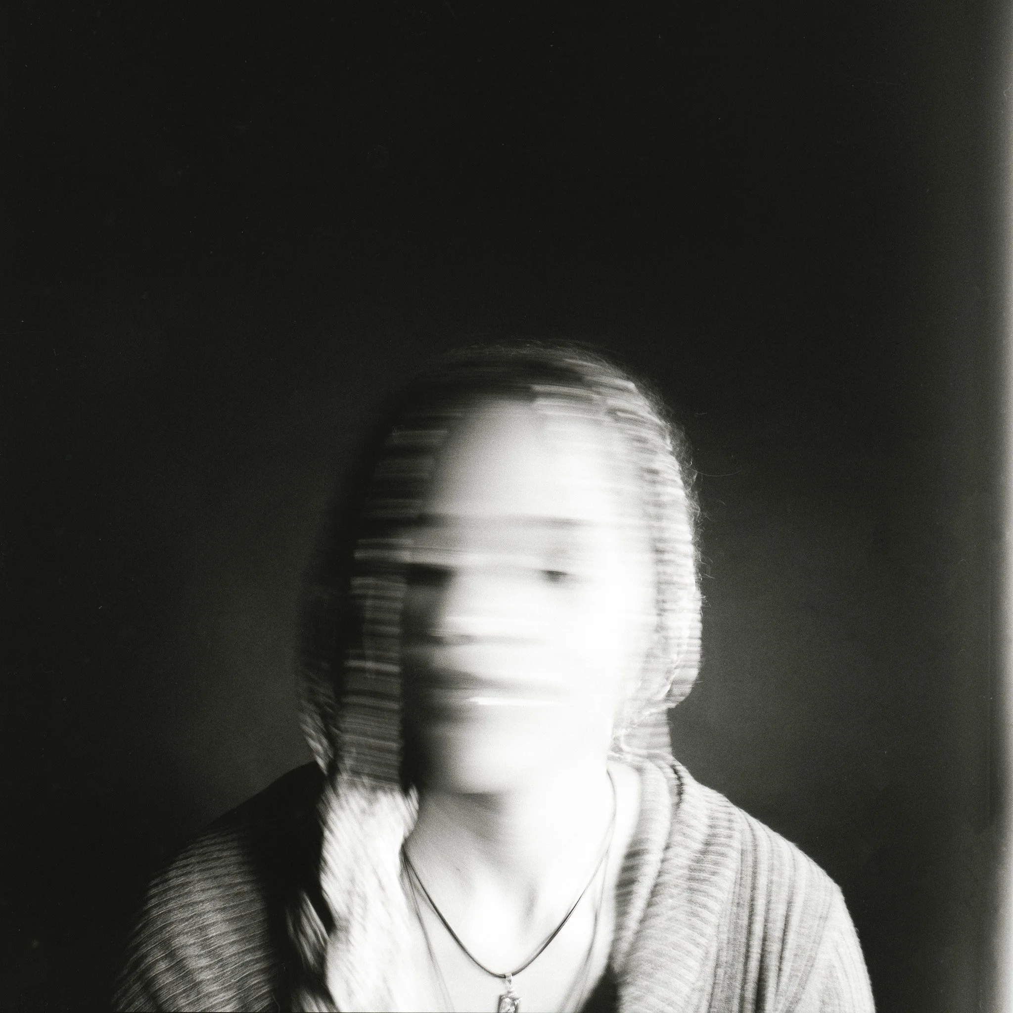 Blurry black-and-white portrait of a woman with shoulder-length hair, wearing a necklace and a striped garment, against a dark background.