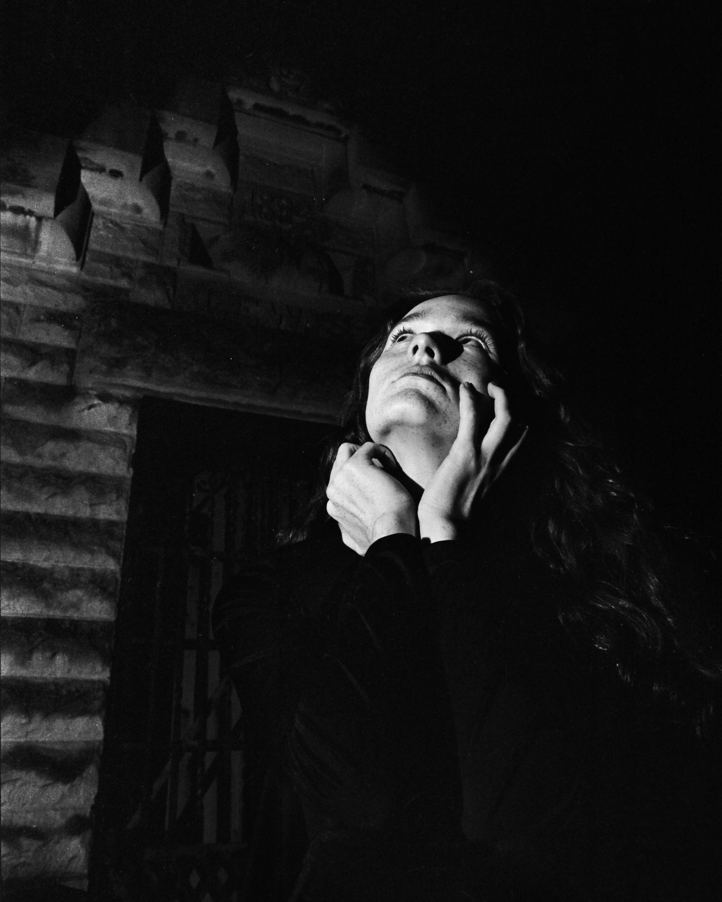 A black and white photo of a woman with wavy hair looking upward, with hands on her chin and cheek, illuminated from below against a dark background.