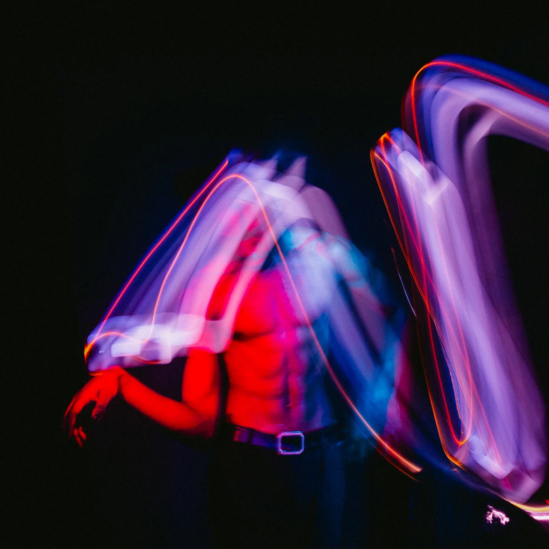 Person in dark clothing moves with long exposure, creating colorful light trails in pink, purple, blue and red against a black background.