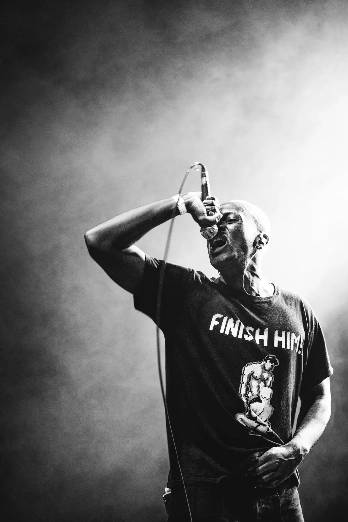 Black and white photo of a man singing passionately into a microphone on stage, wearing a T-shirt with a graphic of a muscular person and the words 'Finish Him!'