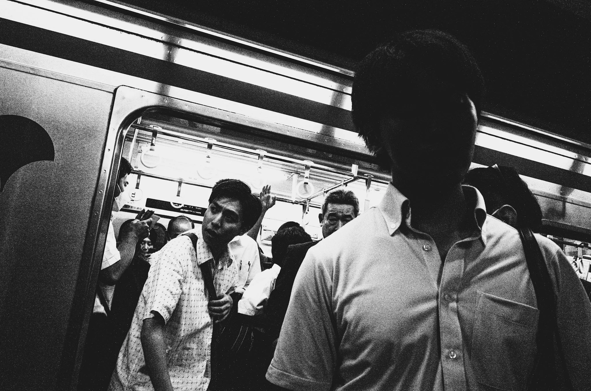 Crowded subway train with passengers standing and holding onto handrails, including a man in a white shirt in the foreground and others of diverse backgrounds in the background.
