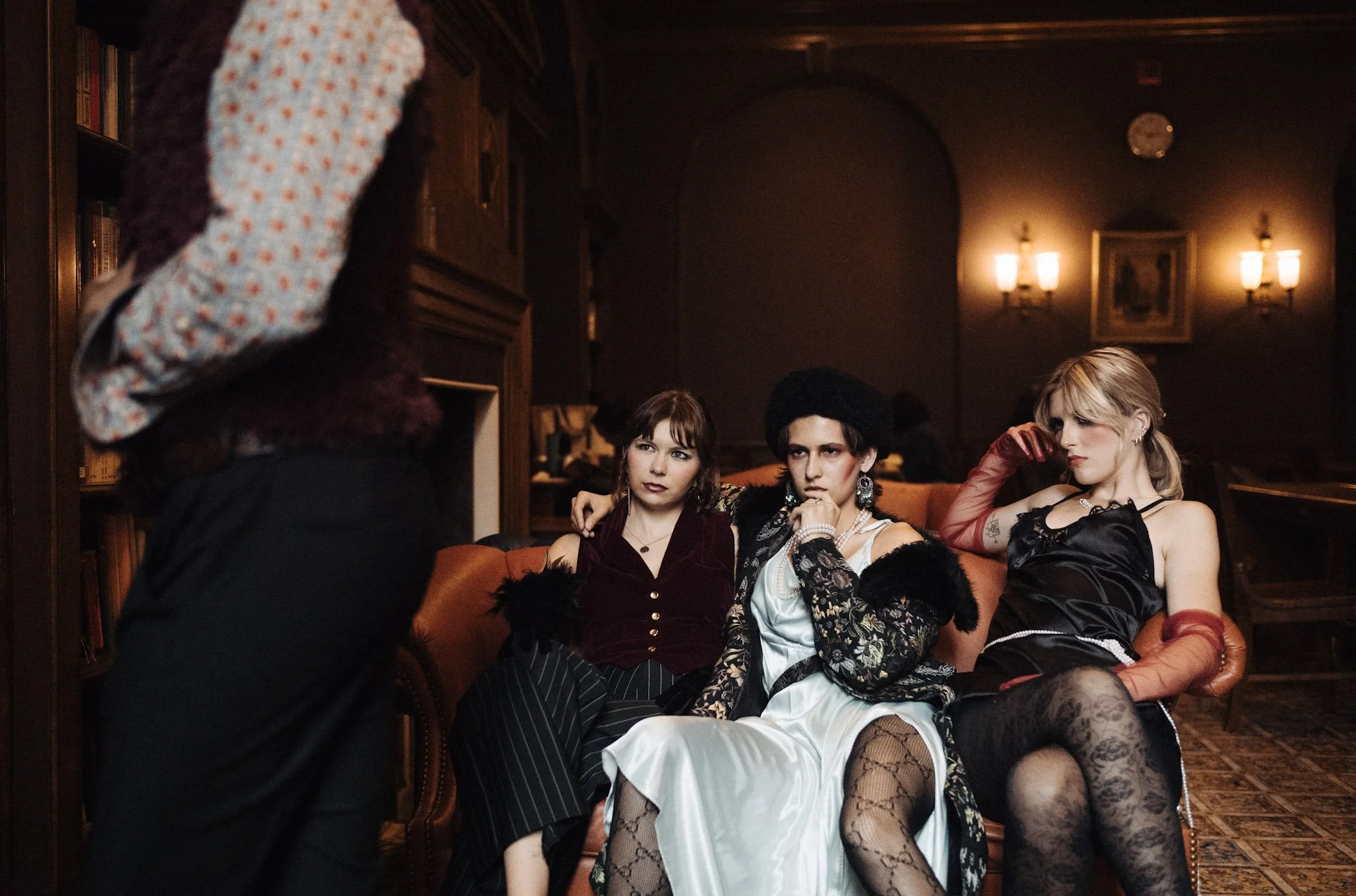 Three women sitting on a vintage sofa in a dimly lit room, dressed in stylish, vintage-inspired clothing, with a person standing in front of them. The room has dark walls, warm lighting, and framed artwork on the wall.