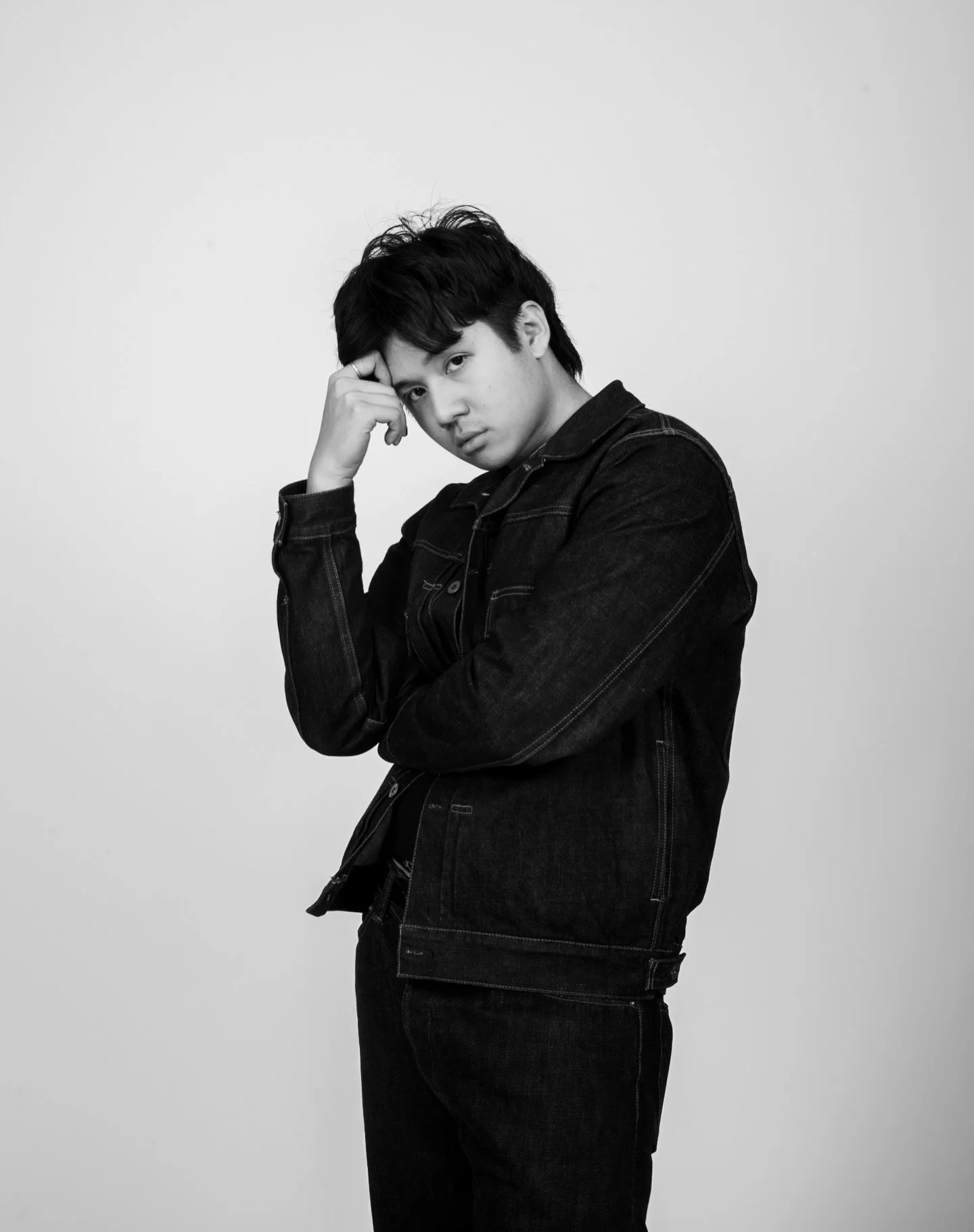 Black and white portrait of a young man with dark hair wearing a denim jacket, standing against a plain background.