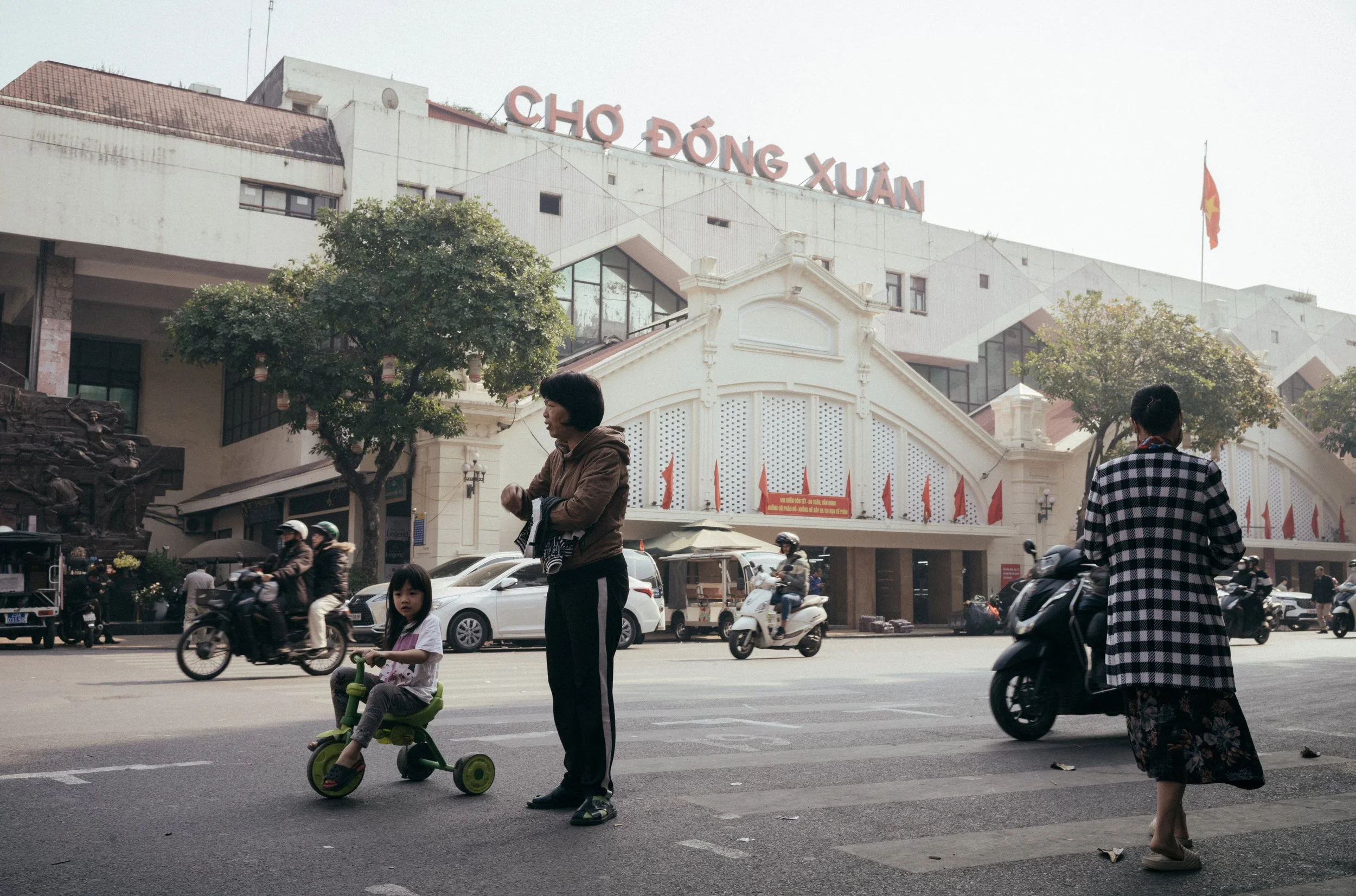 Street scene with a young girl riding a tricycle, woman standing nearby, and a woman on a motorbike in front of a building with signage that reads 'Chợ Đông Xuân'.