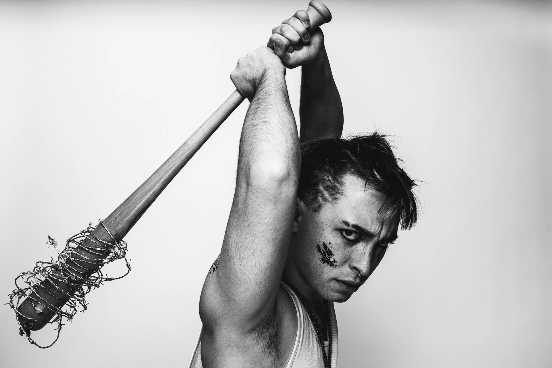Black and white photo of a young man holding an axe over his shoulder, wearing a tank top, with tattoos and a serious expression.